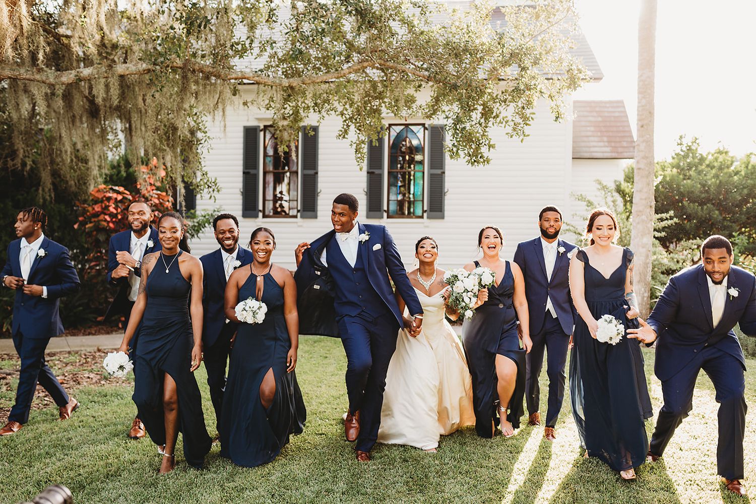 Wedding party in navy blue attire poses outdoors at venue with Spanish moss and white columns in background.