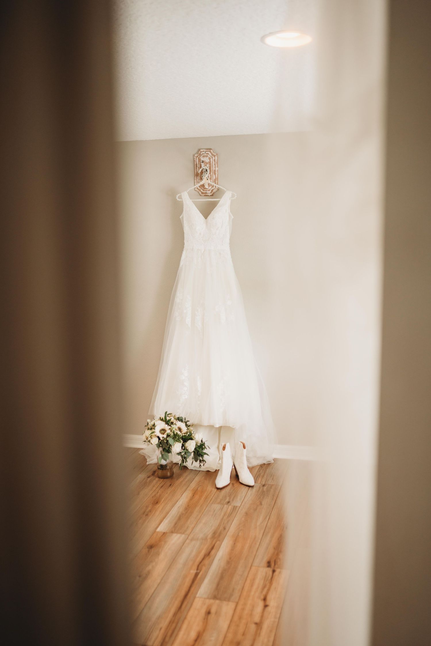 A white wedding dress hangs elegantly on a hanger with white heels and a floral bouquet placed beneath on wooden floors.