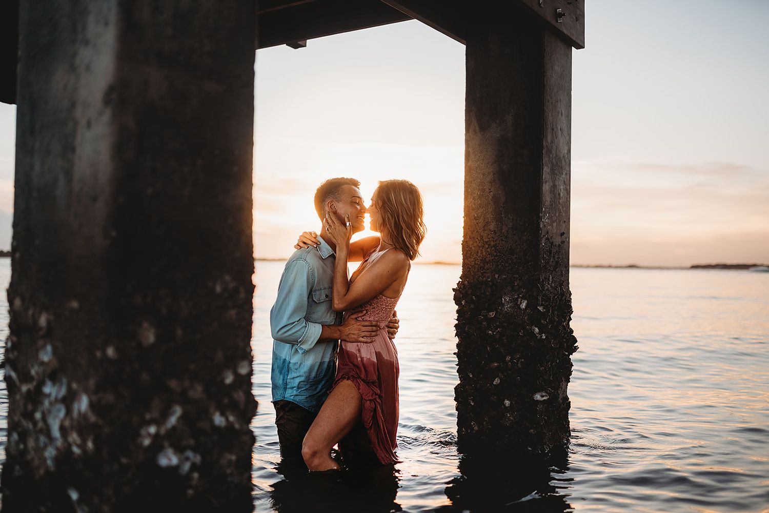 Romantic sunset silhouette beneath pier with calm ocean waters and warm glowing sky in background.
