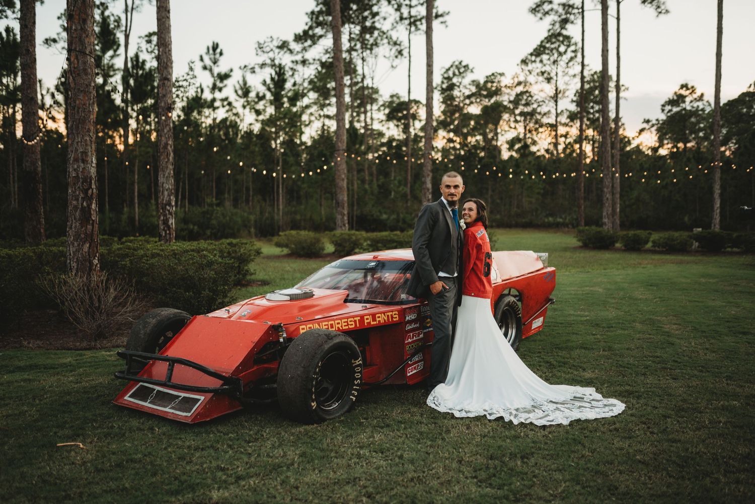 A couple poses with a red formula-style race car on a grassy field surrounded by tall pine trees at sunset.