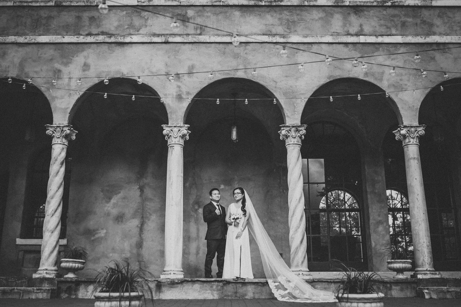 Black and white wedding photo of couple standing under classical stone archway with columns.