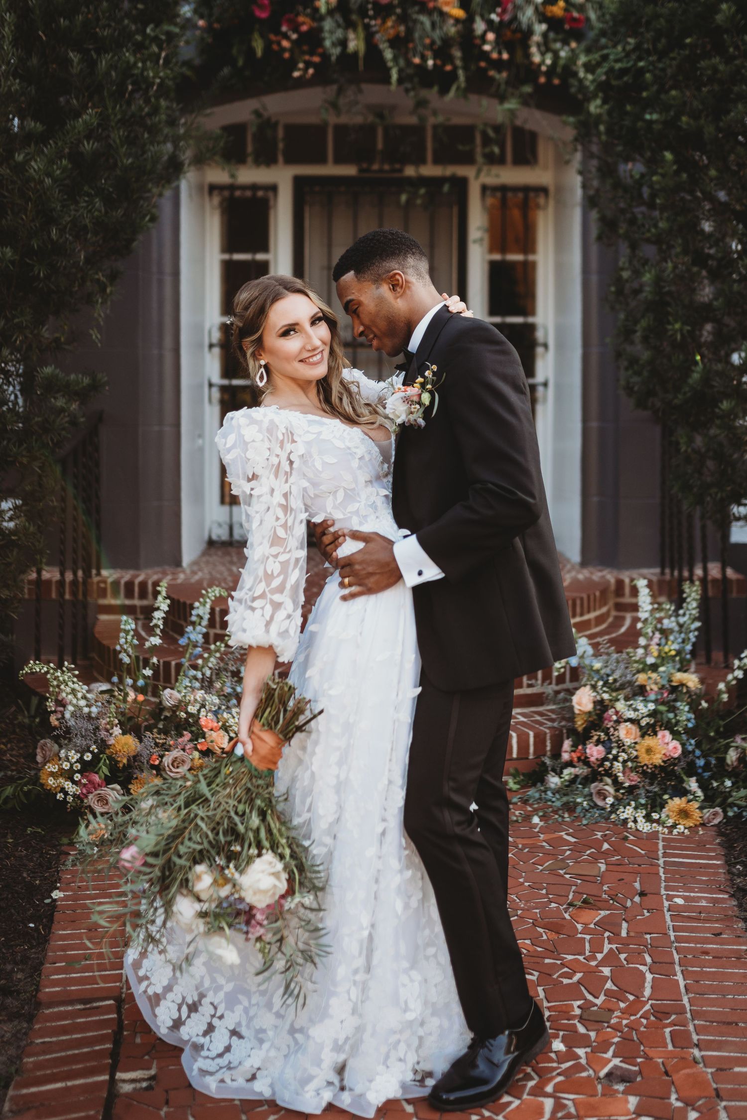 Wedding couple in elegant attire posing together in front of garden doorway with floral arch.