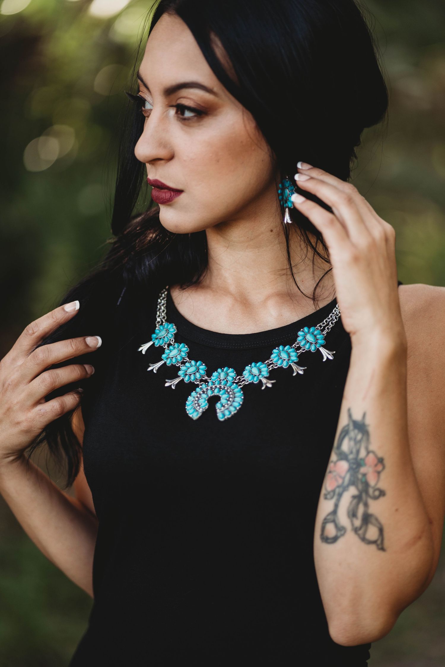 A person wearing a black dress with a turquoise statement necklace and visible arm tattoo touches their dark hair.