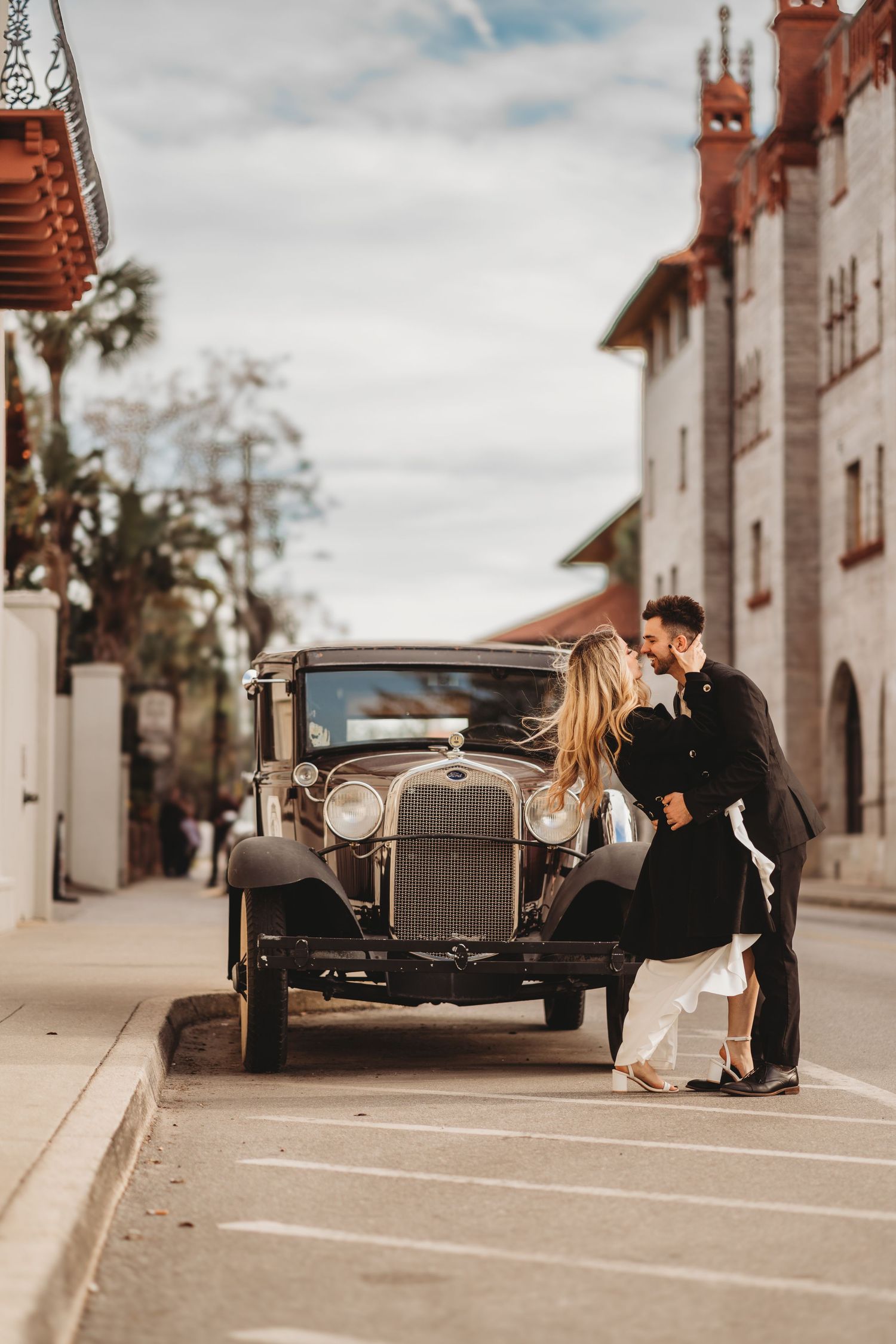 A romantic vintage scene with a couple embracing next to a classic 1920s car on a historic Spanish-style street.