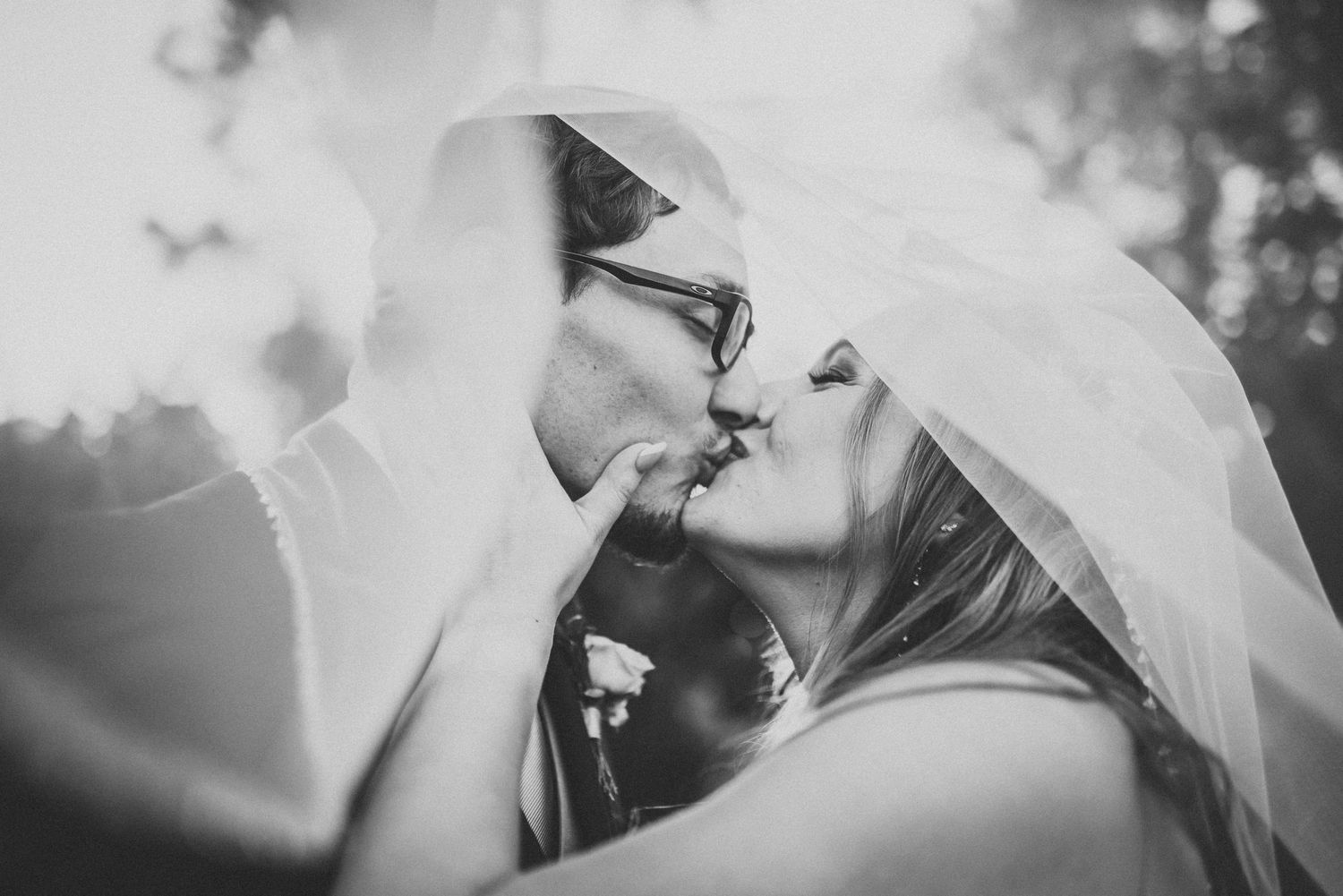 Black and white wedding photograph showing a romantic kiss between newlyweds under a soft bridal veil.