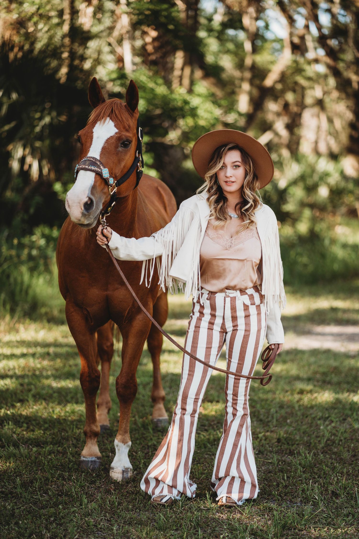 A stylish woman in striped bell bottoms and brown hat stands with a chestnut horse on a wooded trail.