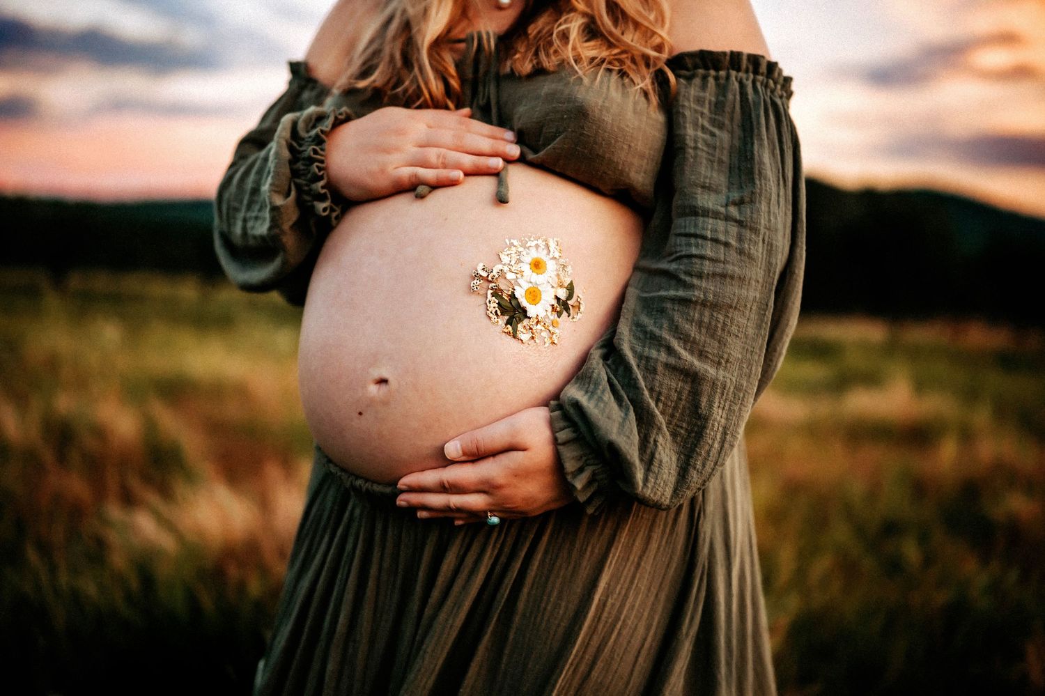 Pregnant woman in olive green dress cradling bare baby bump with daisy flowers at golden hour sunset.