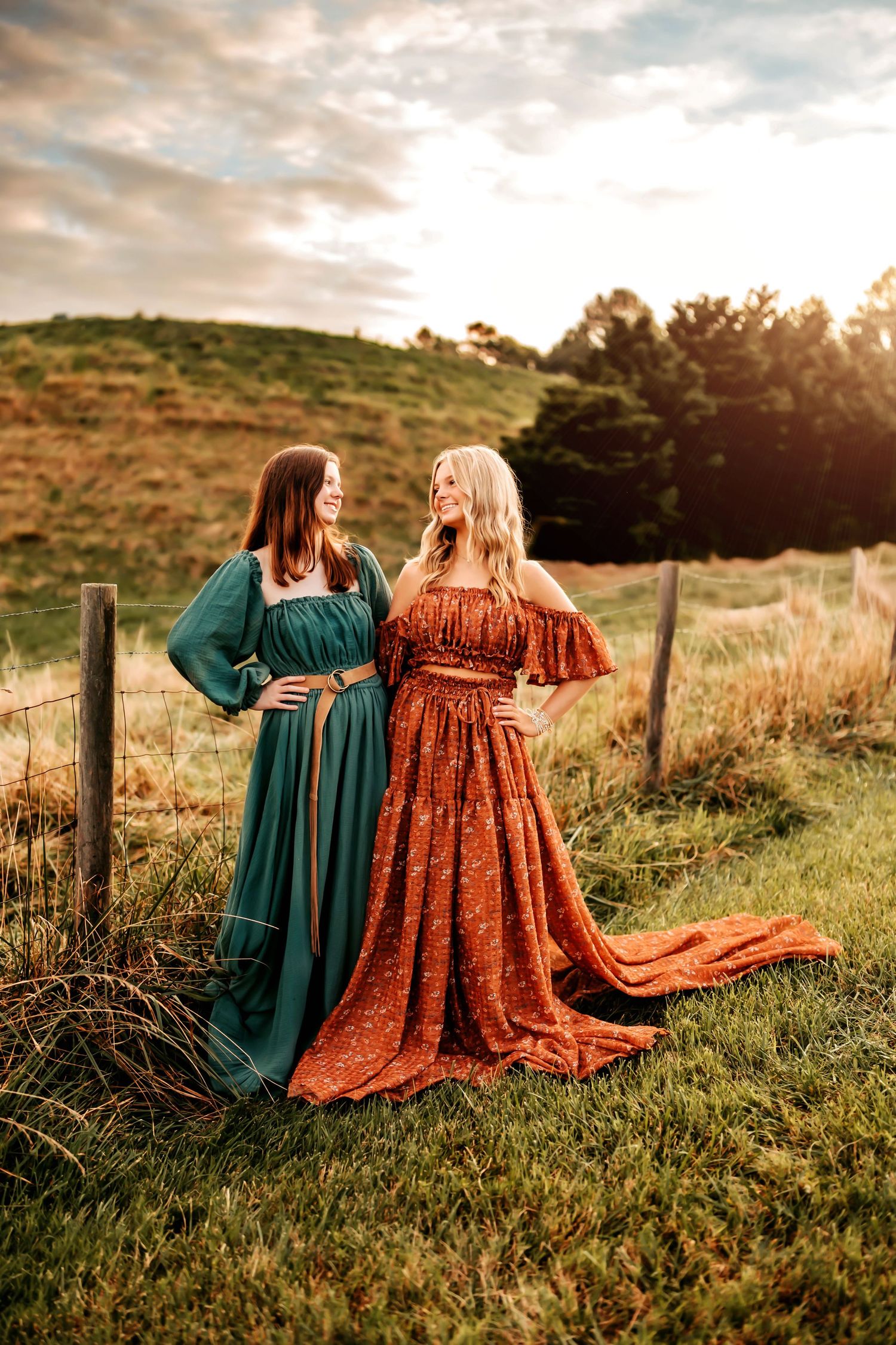 Two people in flowing dresses sit together on a grassy hillside at sunset enjoying the peaceful scenery.