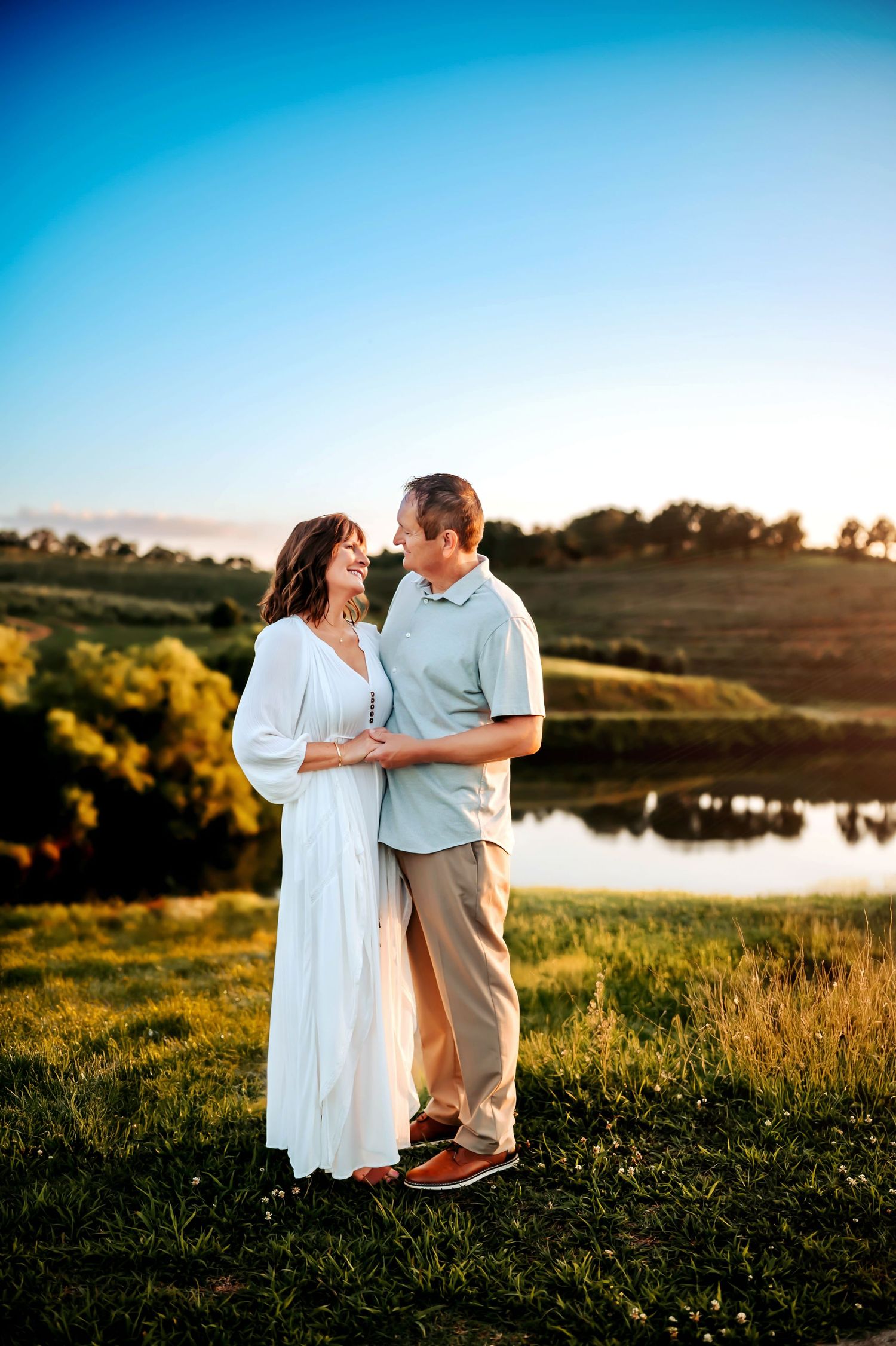 Couple embracing and smiling at each other by a reflective pond at golden hour sunset.