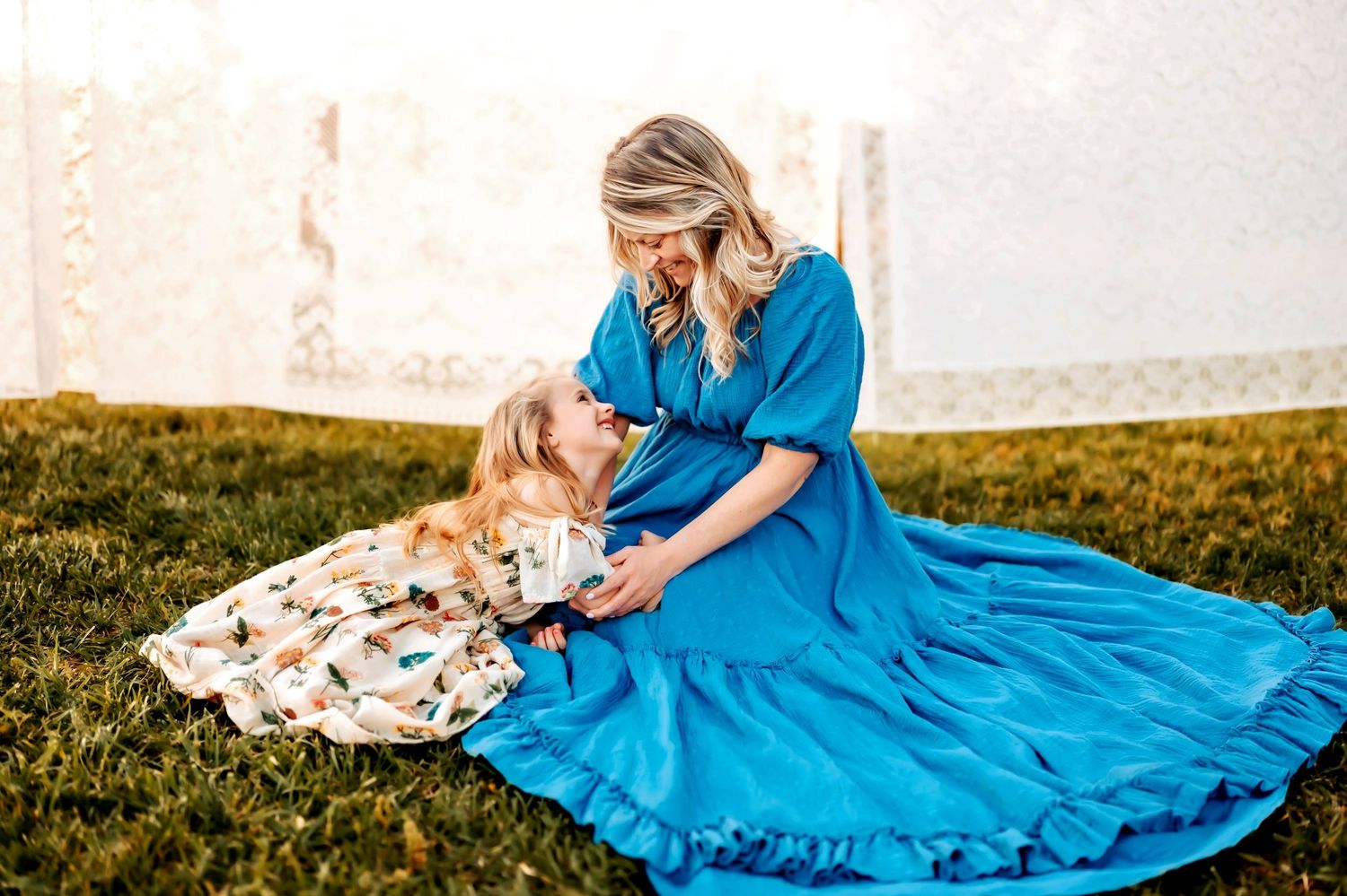 A mother and daughter in matching floral and blue dresses share a tender moment while sitting in a grassy field.