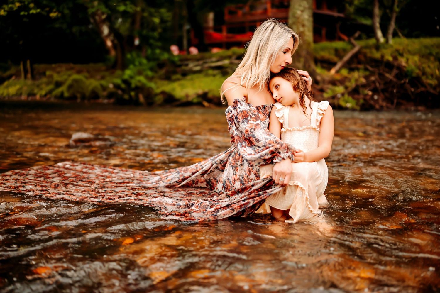 A tender moment captured in a flowing floral dress as two figures embrace while sitting in shallow creek waters at sunset.