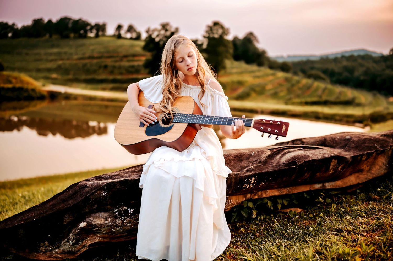 Someone in a flowing white dress plays acoustic guitar while seated on a log by a lake at sunset.