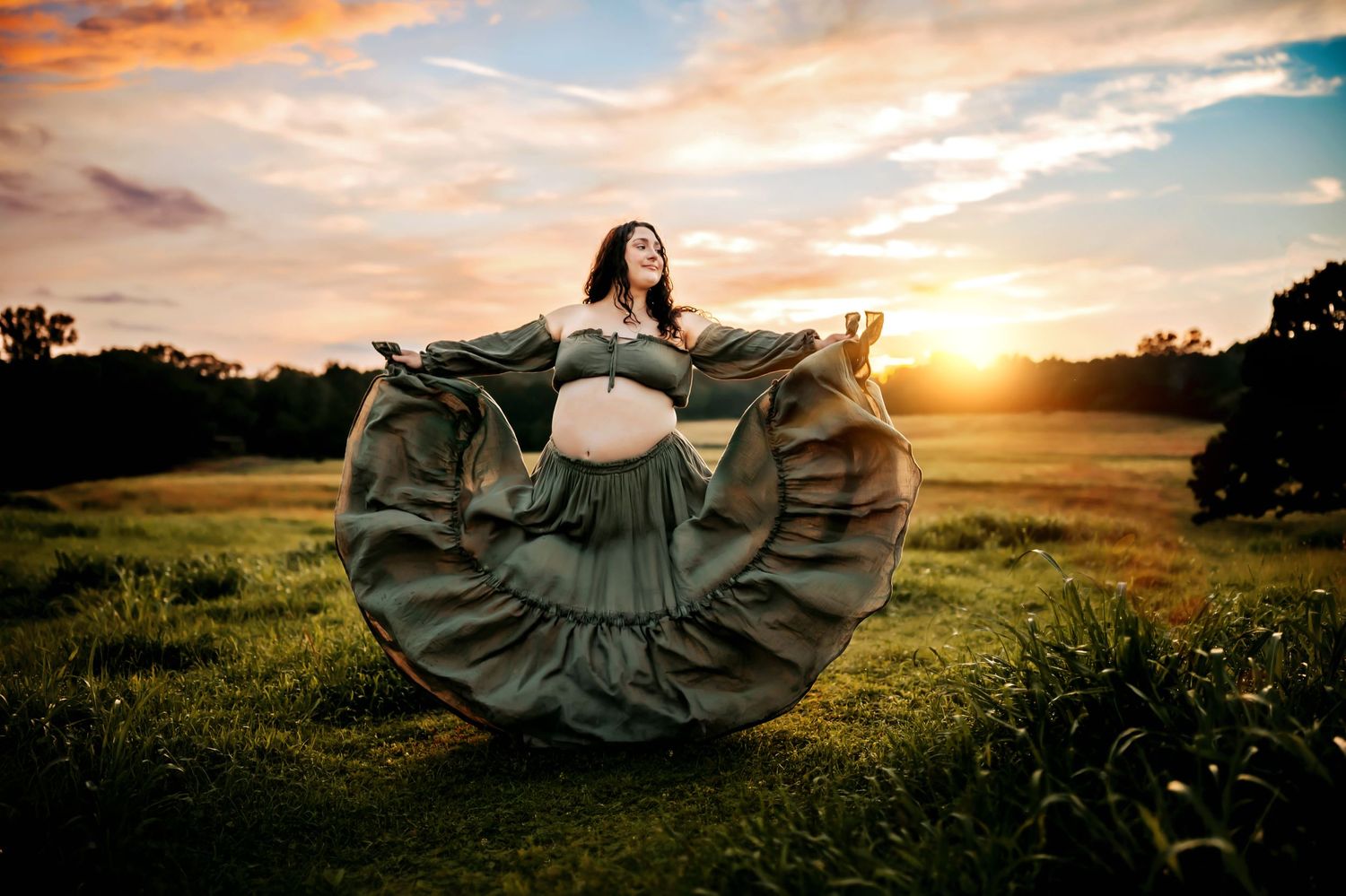 Pregnant woman in flowing gray gown poses at golden sunset in green field, maternity photography.