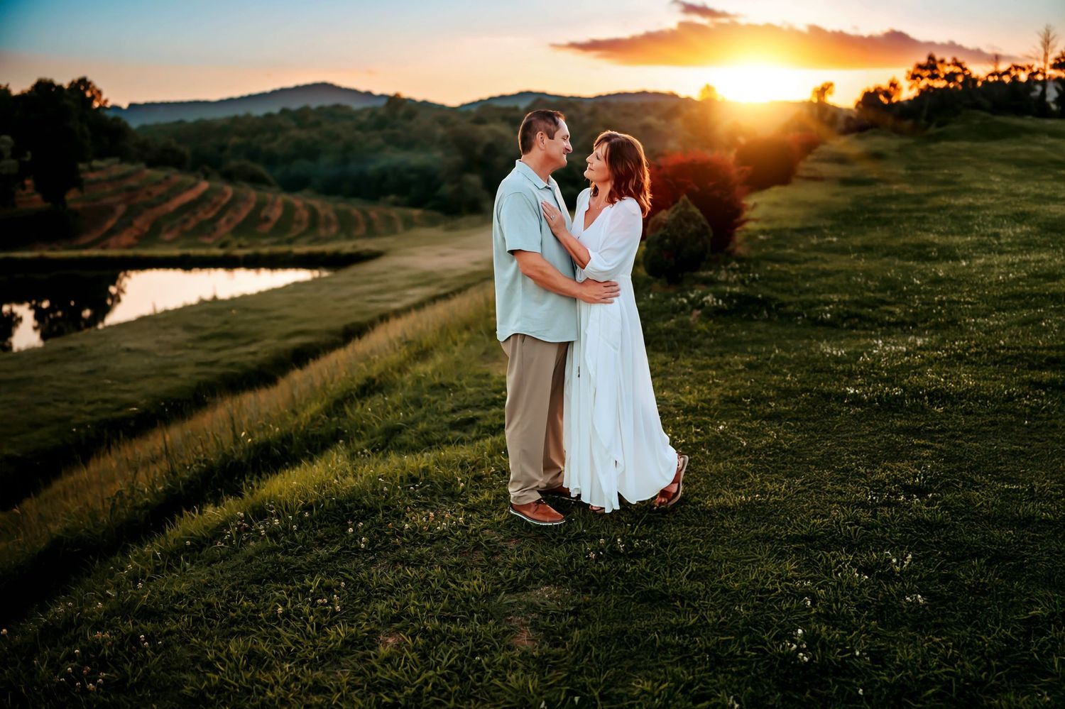 Couple embracing at golden sunset in lush green vineyard landscape with pond and rolling hills.