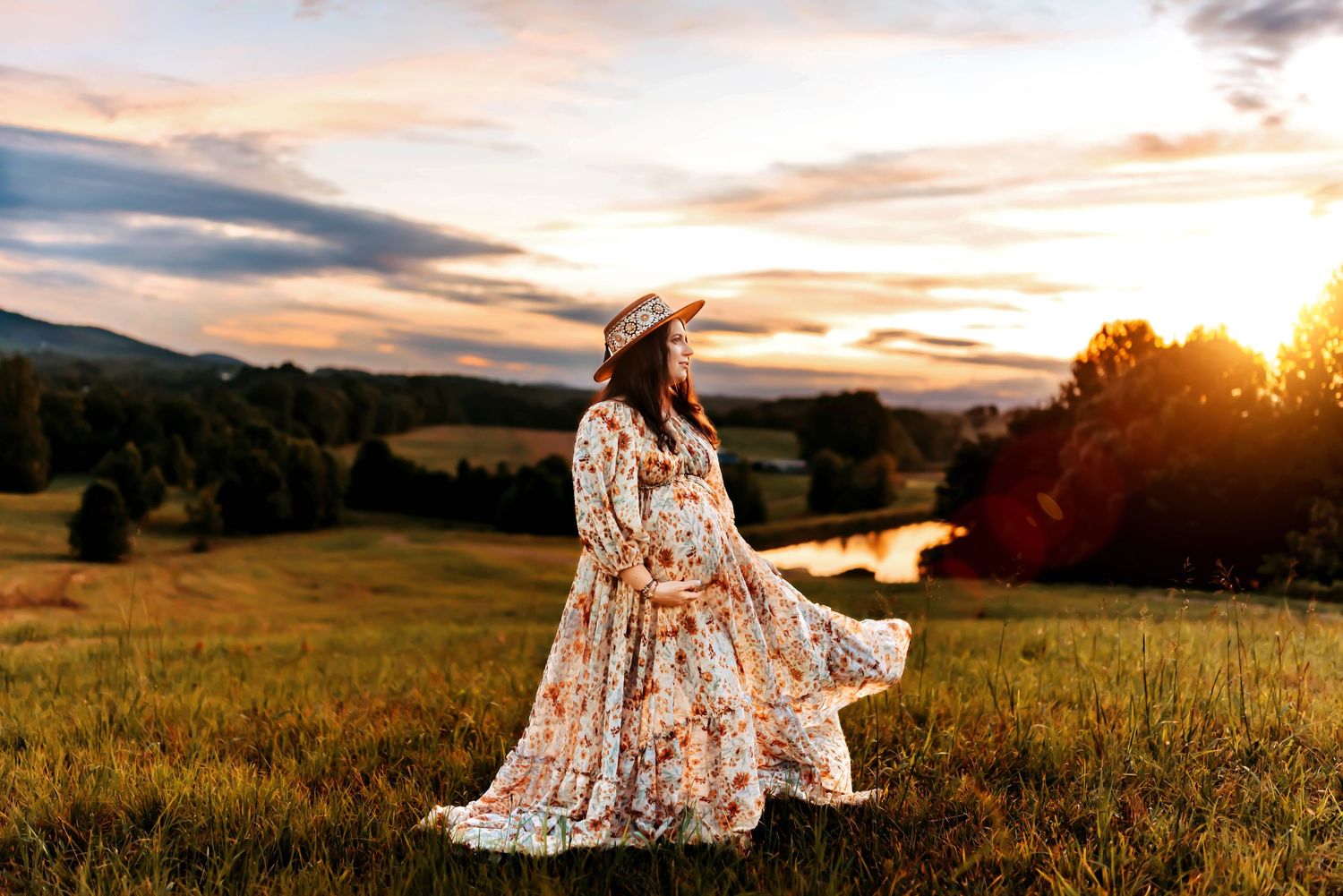 Pregnant woman in floral dress and hat standing in golden sunset meadow with mountains and river in background.