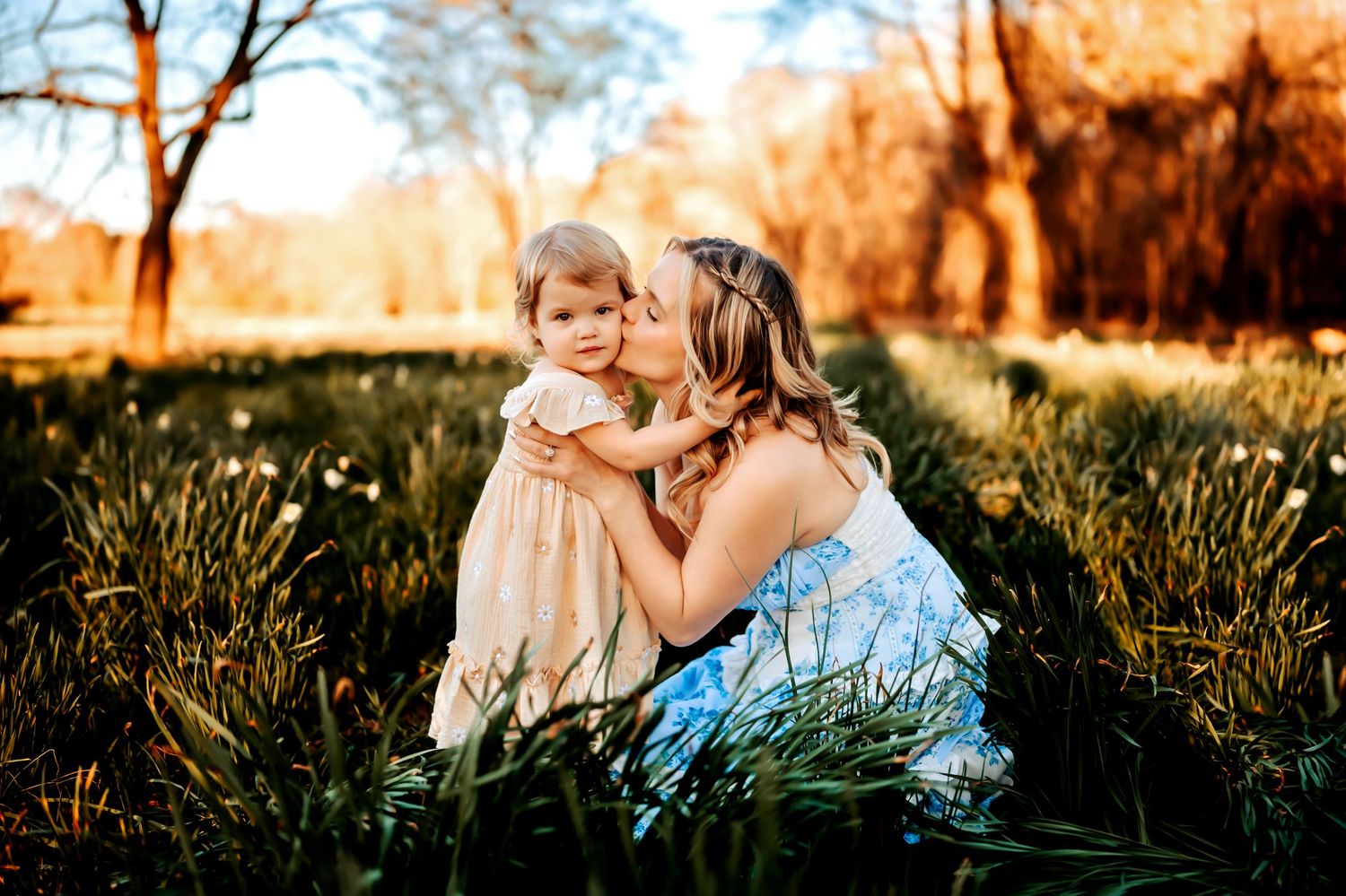 Two people share a tender moment in a grassy field during golden hour with sunlit trees in the background.