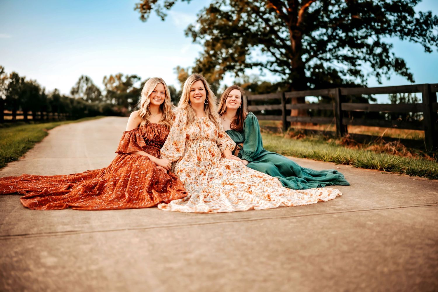 A group of people in elegant dresses sit together on a rustic dirt road at sunset creating a dreamy countryside scene.