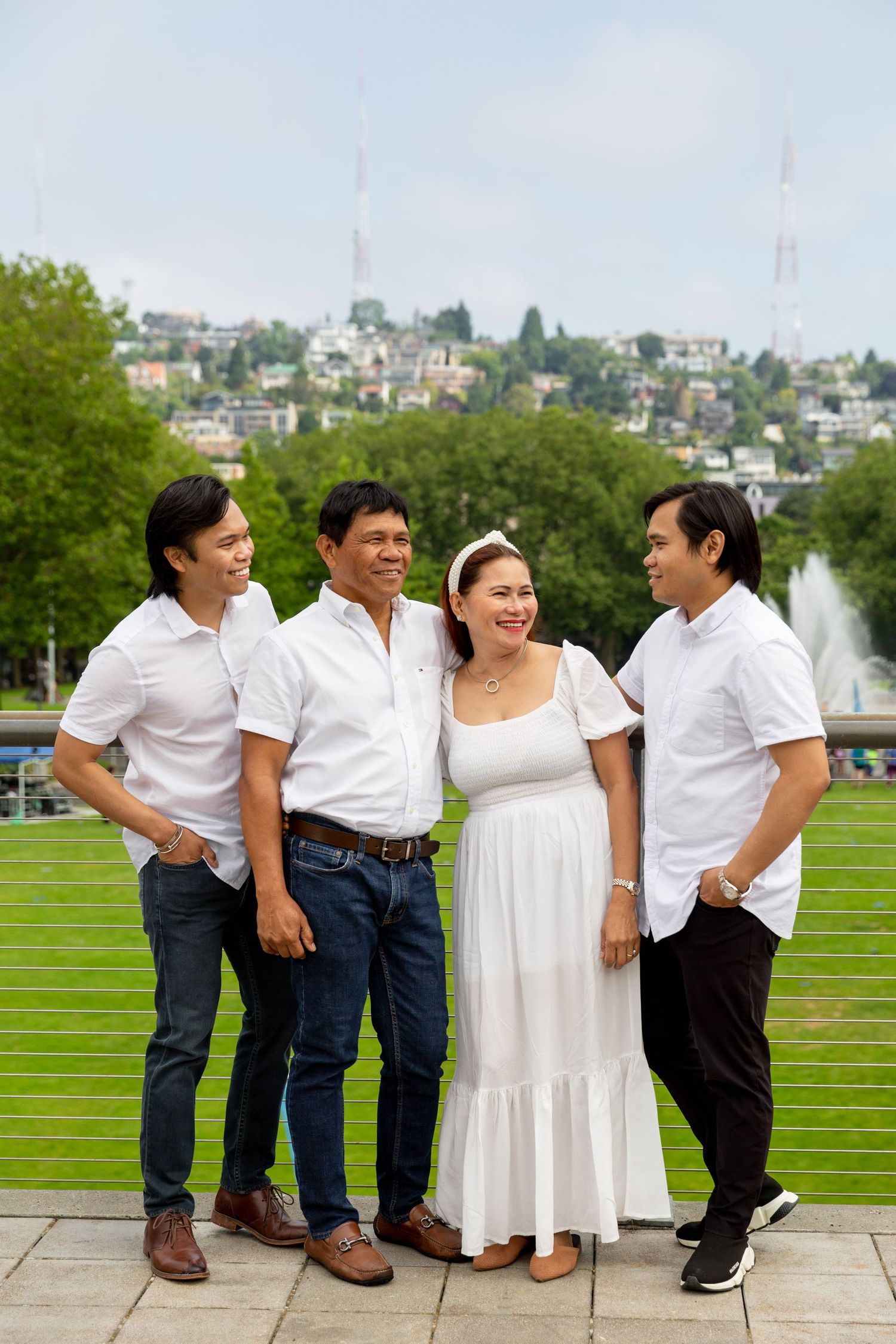 Four people in white outfits pose together in front of a green park landscape with a fountain and city skyline.