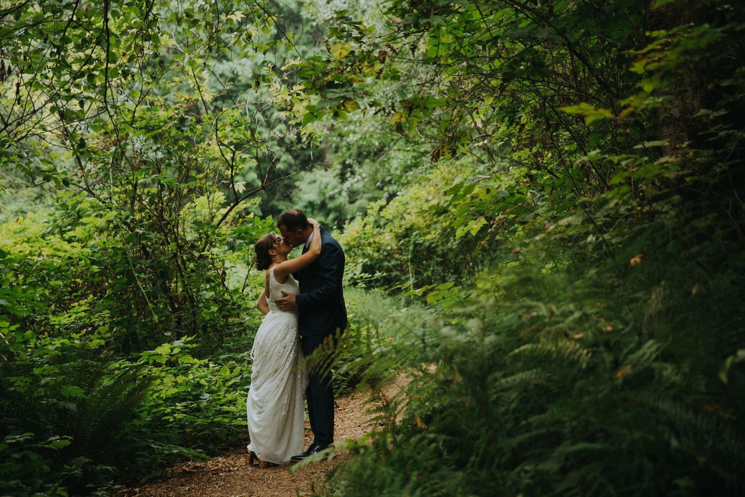 A couple embraces on a forest path surrounded by lush green foliage and natural sunlight filtering through trees.