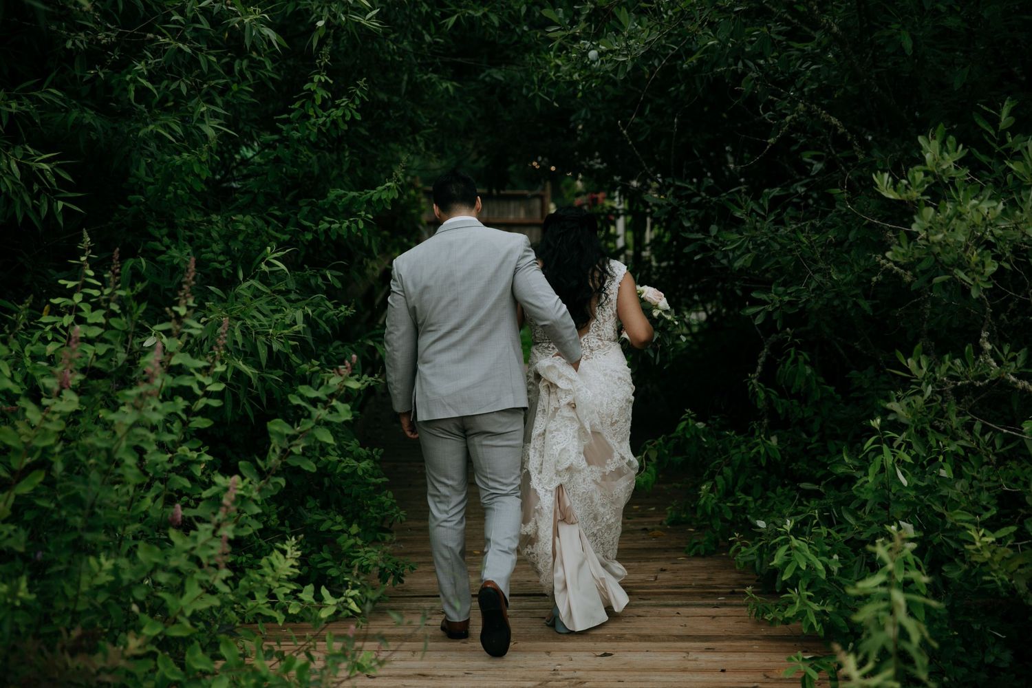 A couple in wedding attire walks along a wooden boardwalk surrounded by lush green forest foliage.