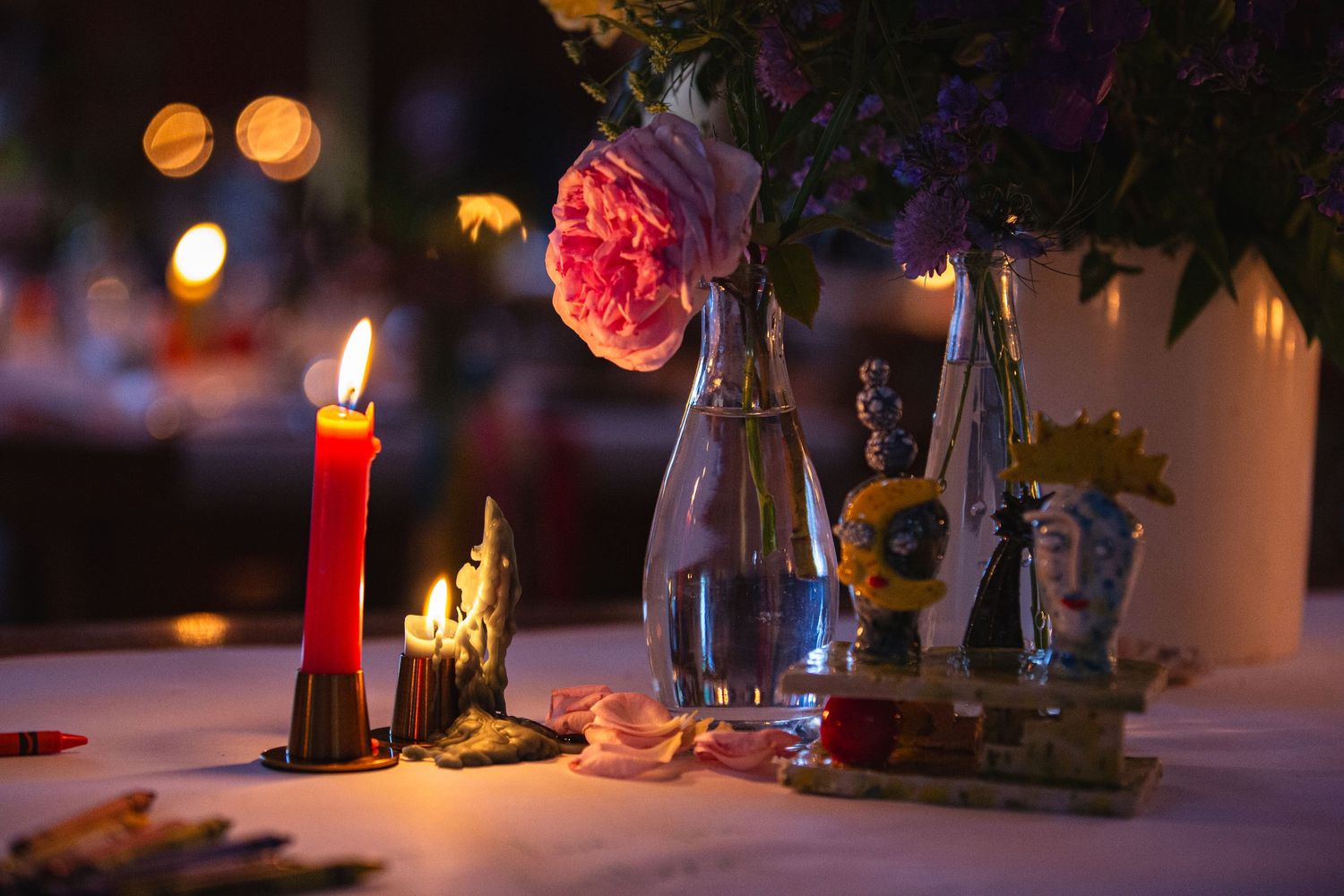 Romantic table setting with lit red candle, pink rose in glass vase, and glowing bokeh lights in background.