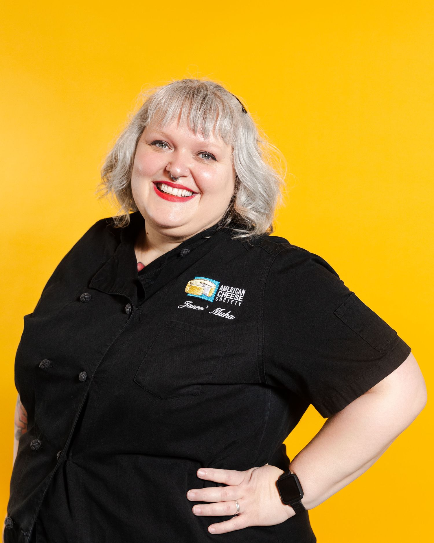 A cheerful person wearing a black work uniform poses with hand on hip against a bright yellow background.