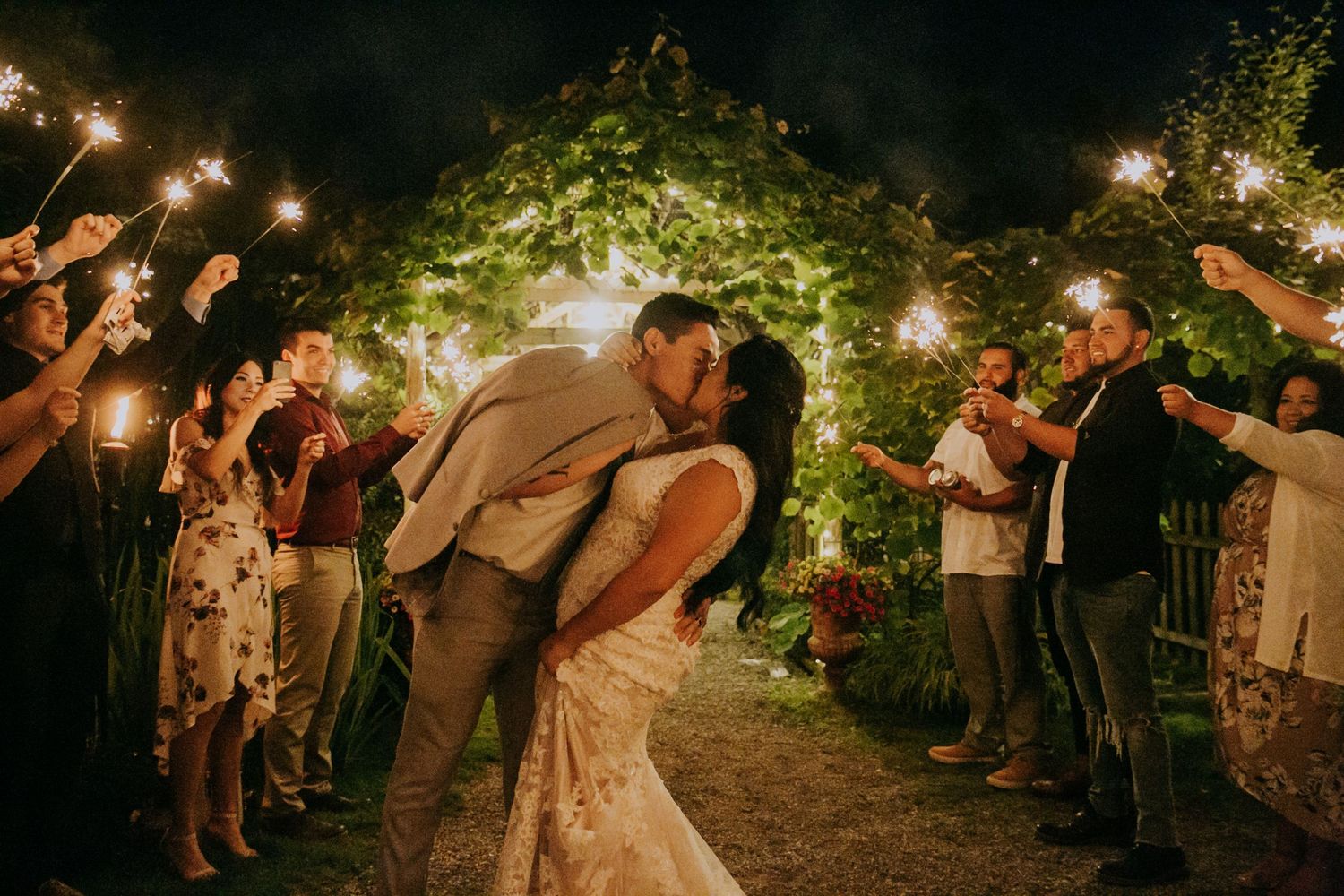 Couple sharing romantic kiss during evening sparkler exit at garden wedding ceremony with guests holding sparklers.