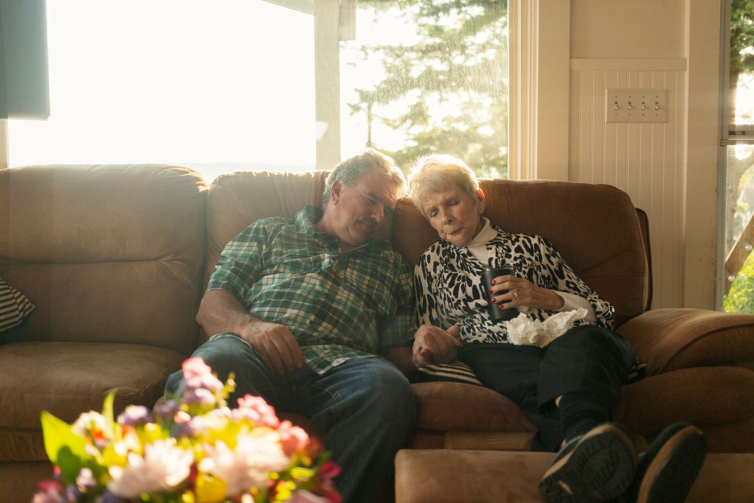 Two elderly people share a tender moment together on a brown couch with flowers in the foreground.