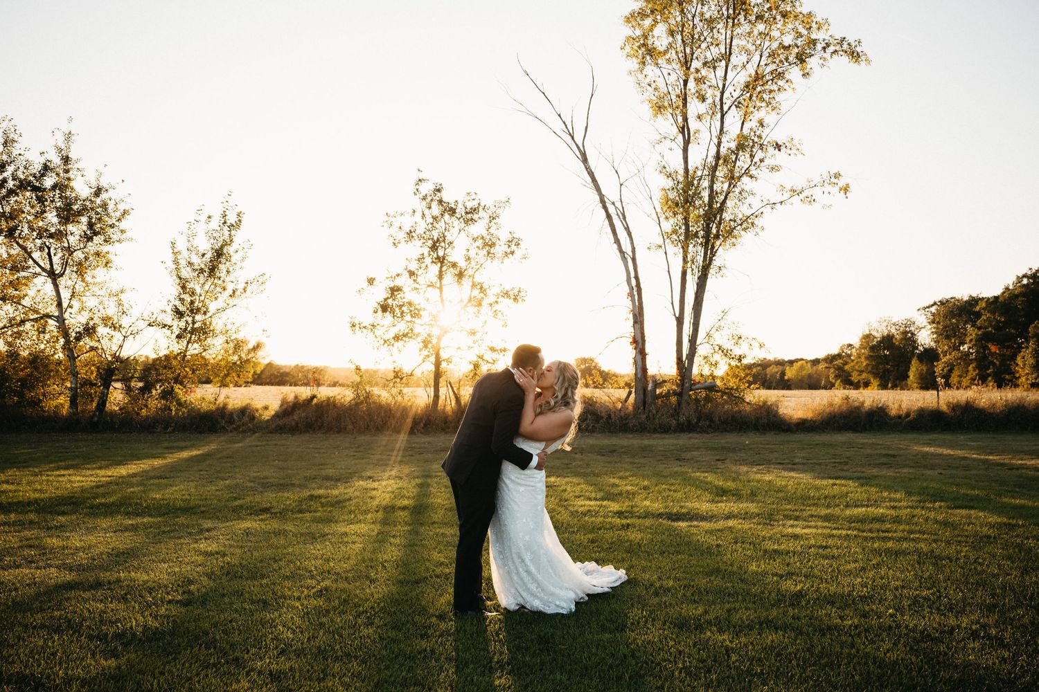 A romantic sunset wedding photo of a couple embracing on a grassy field with golden light filtering through trees.