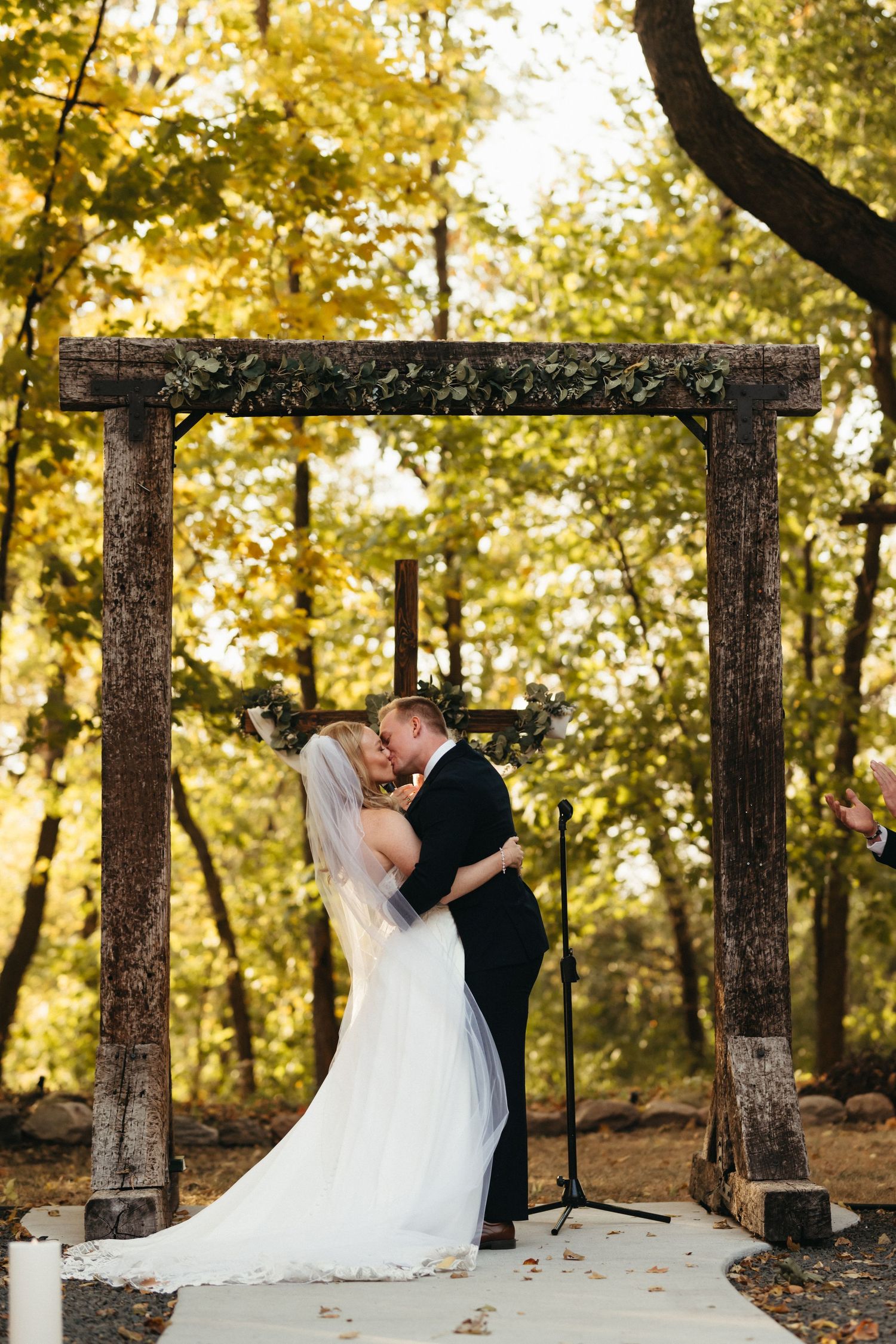 A couple shares their first kiss under a rustic wooden arch surrounded by golden autumn foliage.