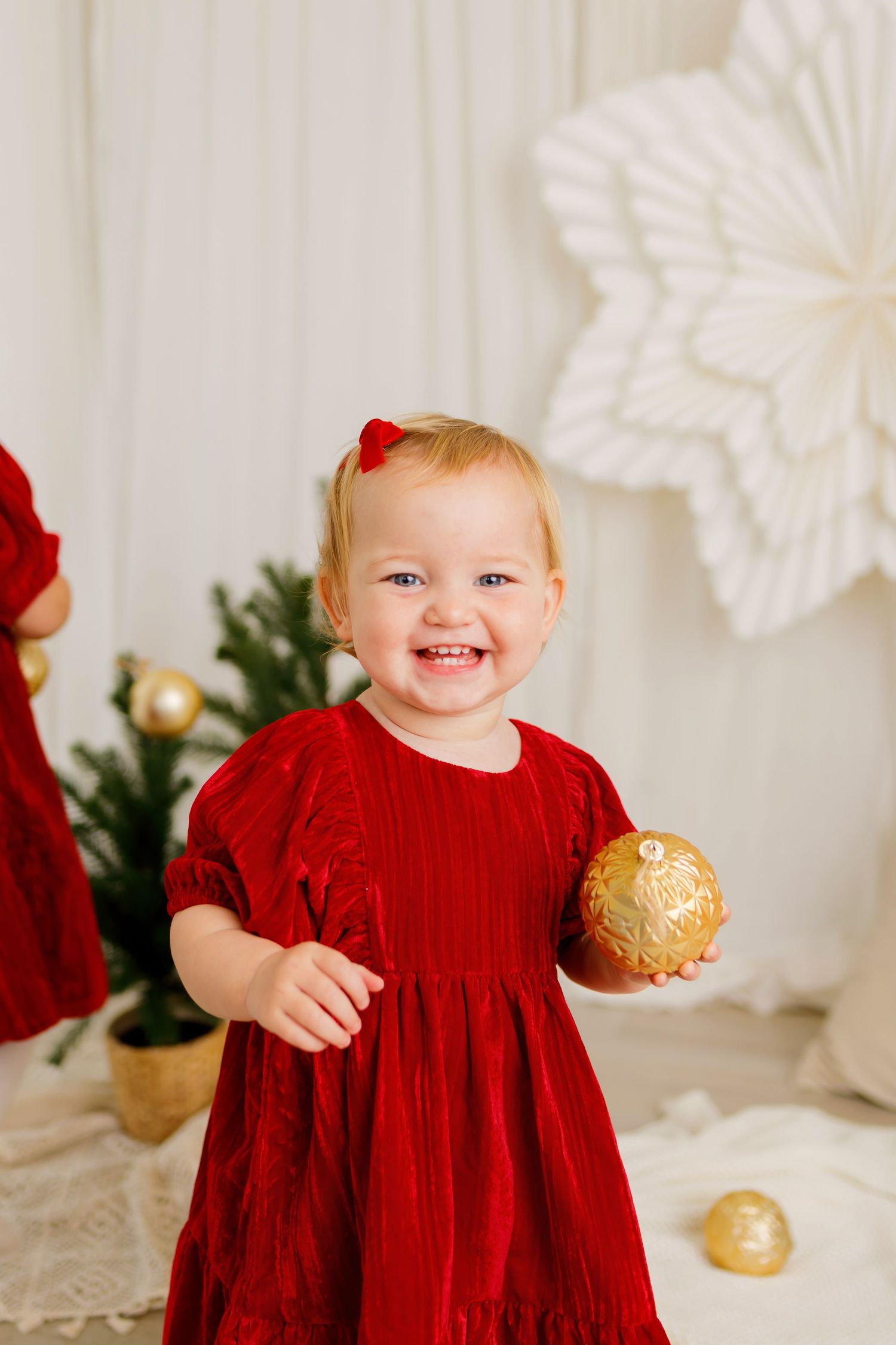 Smiling toddler girl in red velvet dress holding gold Christmas ornament near decorated tree.