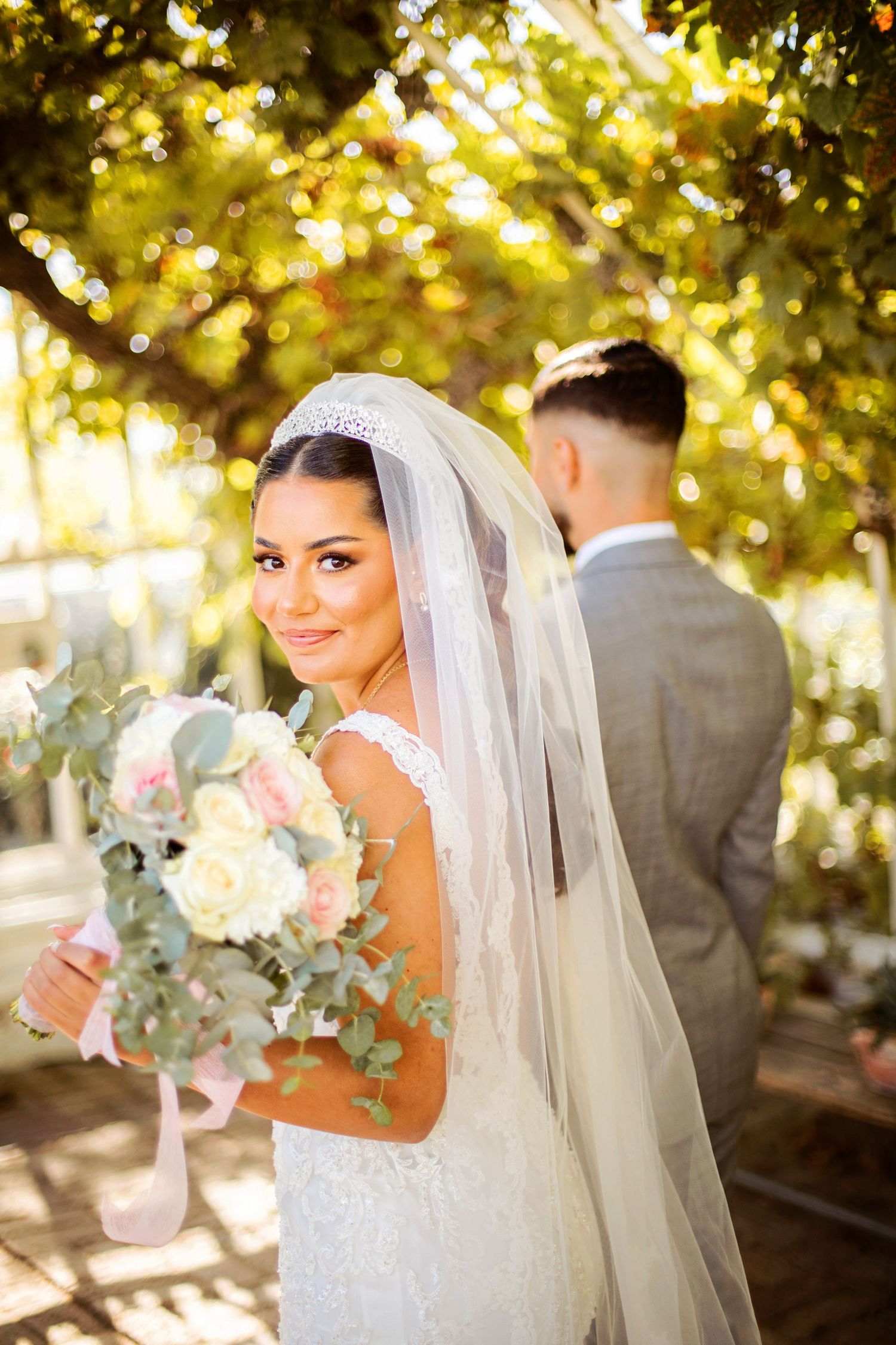 Bride in white lace gown and veil holds floral bouquet, smiling outdoors with groom in gray suit behind her.