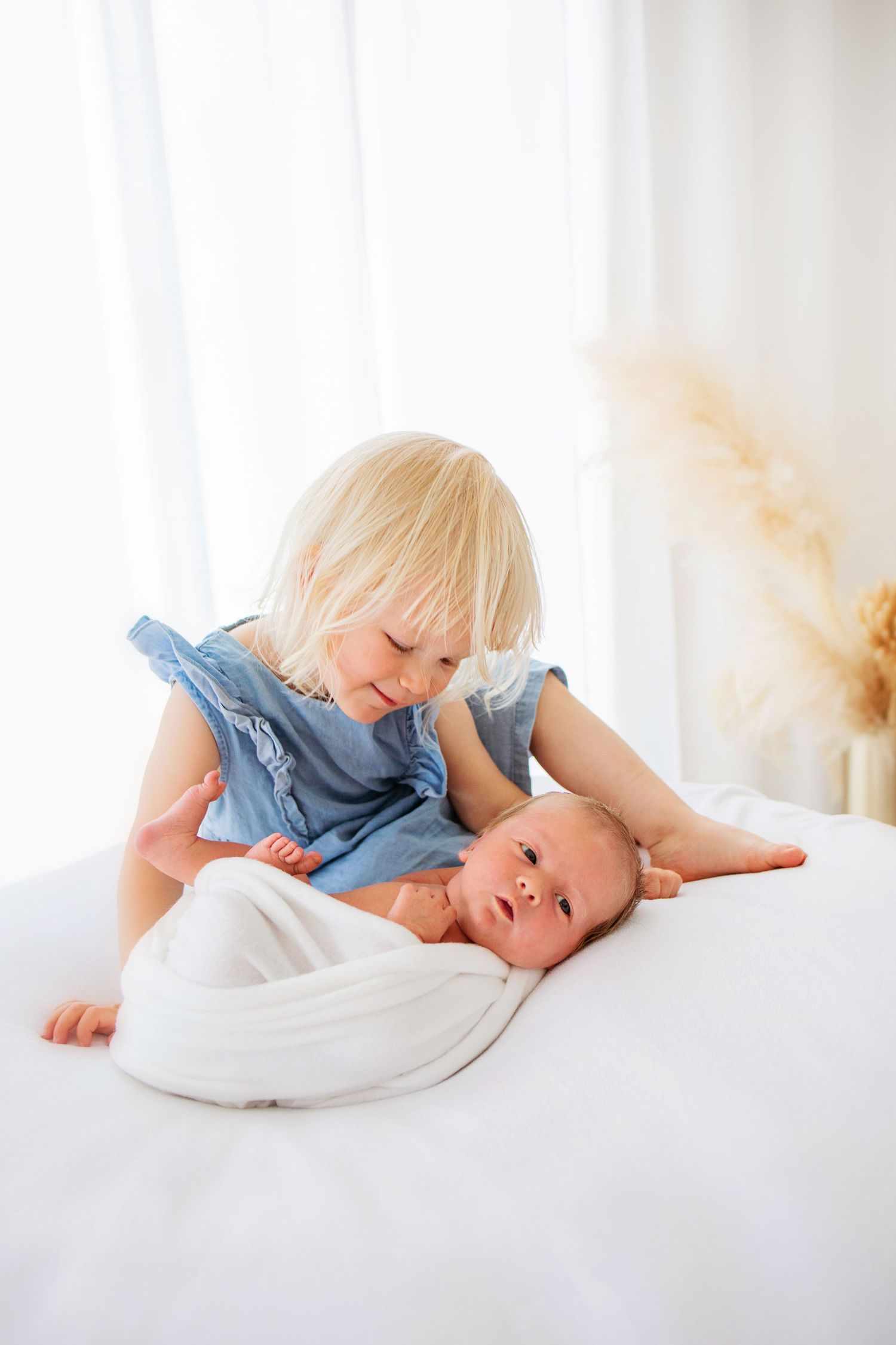 Two young children in white and blue outfits playing together on a bright white bed.