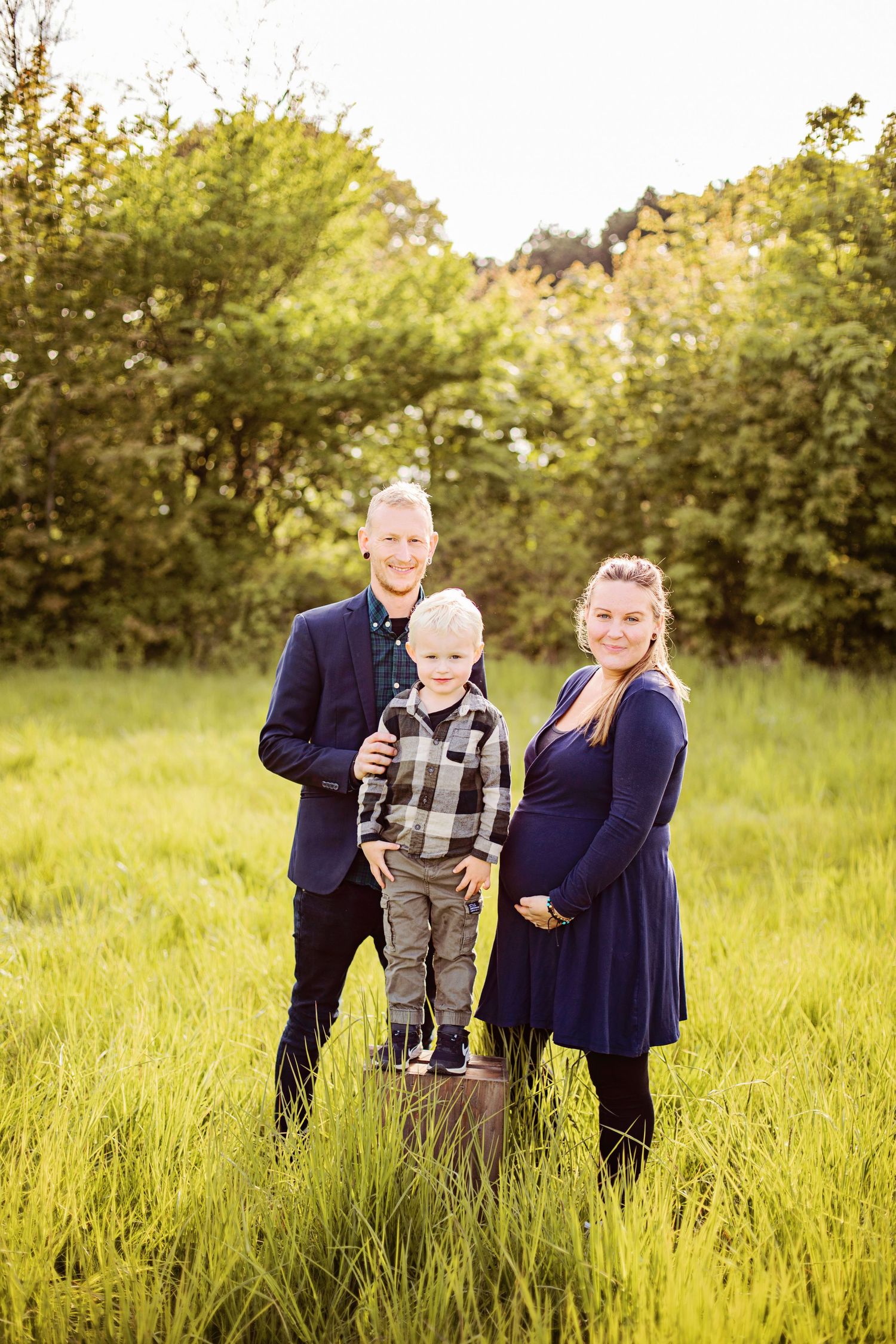 Family of three posing in a lush green meadow, parents in navy blue outfits with young blonde boy between them.