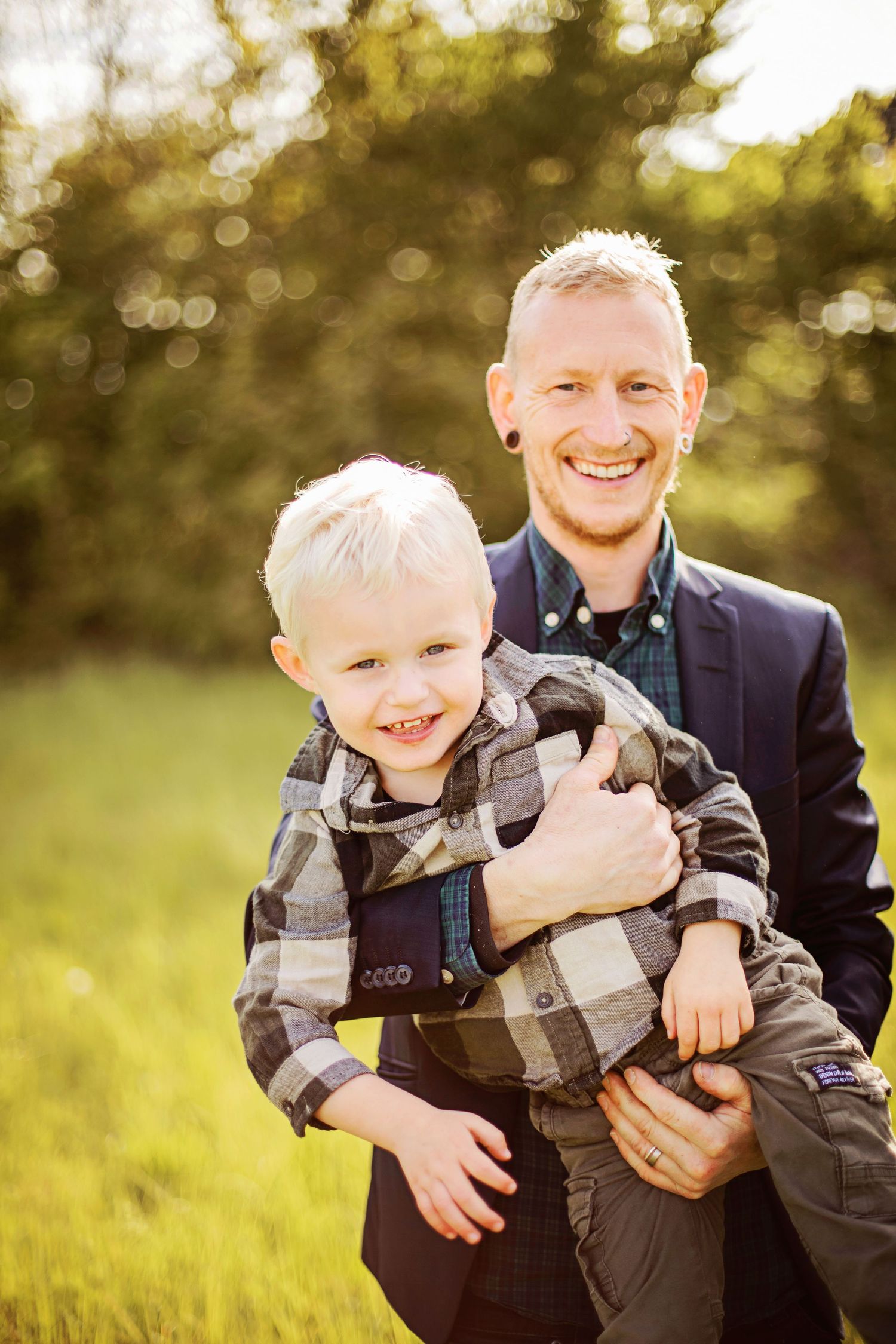 Smiling man in navy blazer holding happy blonde toddler boy in plaid shirt outdoors in sunny green field.