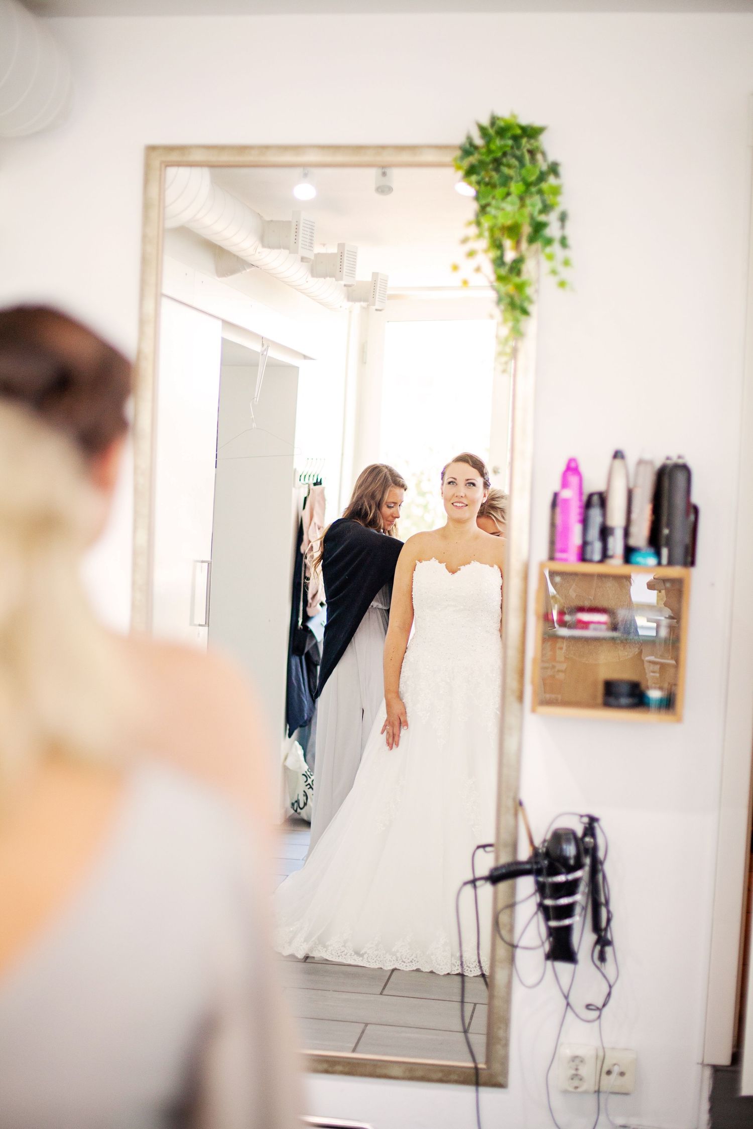 Bride in white strapless wedding gown being helped by stylist, reflected in large mirror in bright bridal salon.