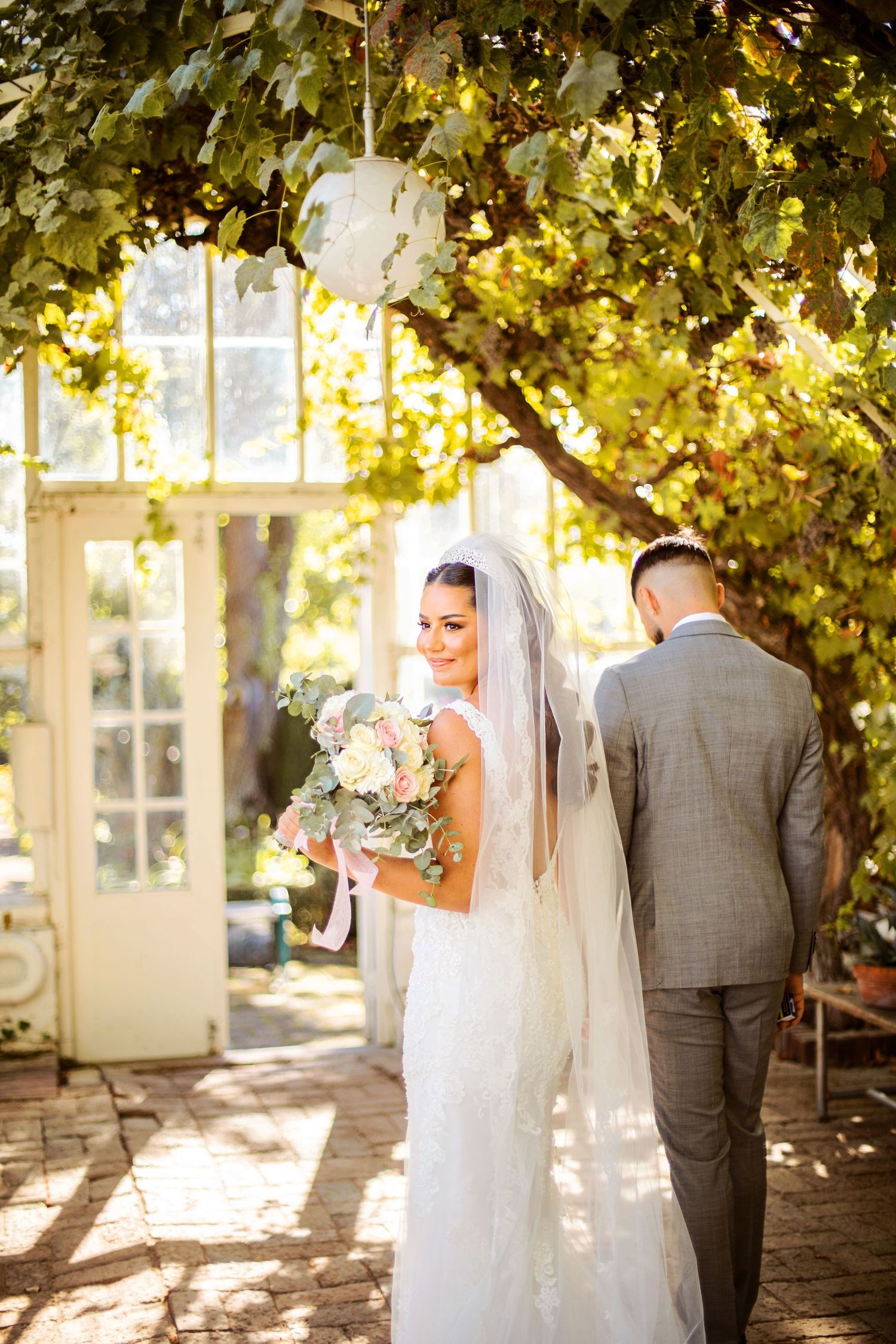 Bride in white gown and veil holds bouquet beside groom in gray suit under sunlit green arbor near vintage doors.