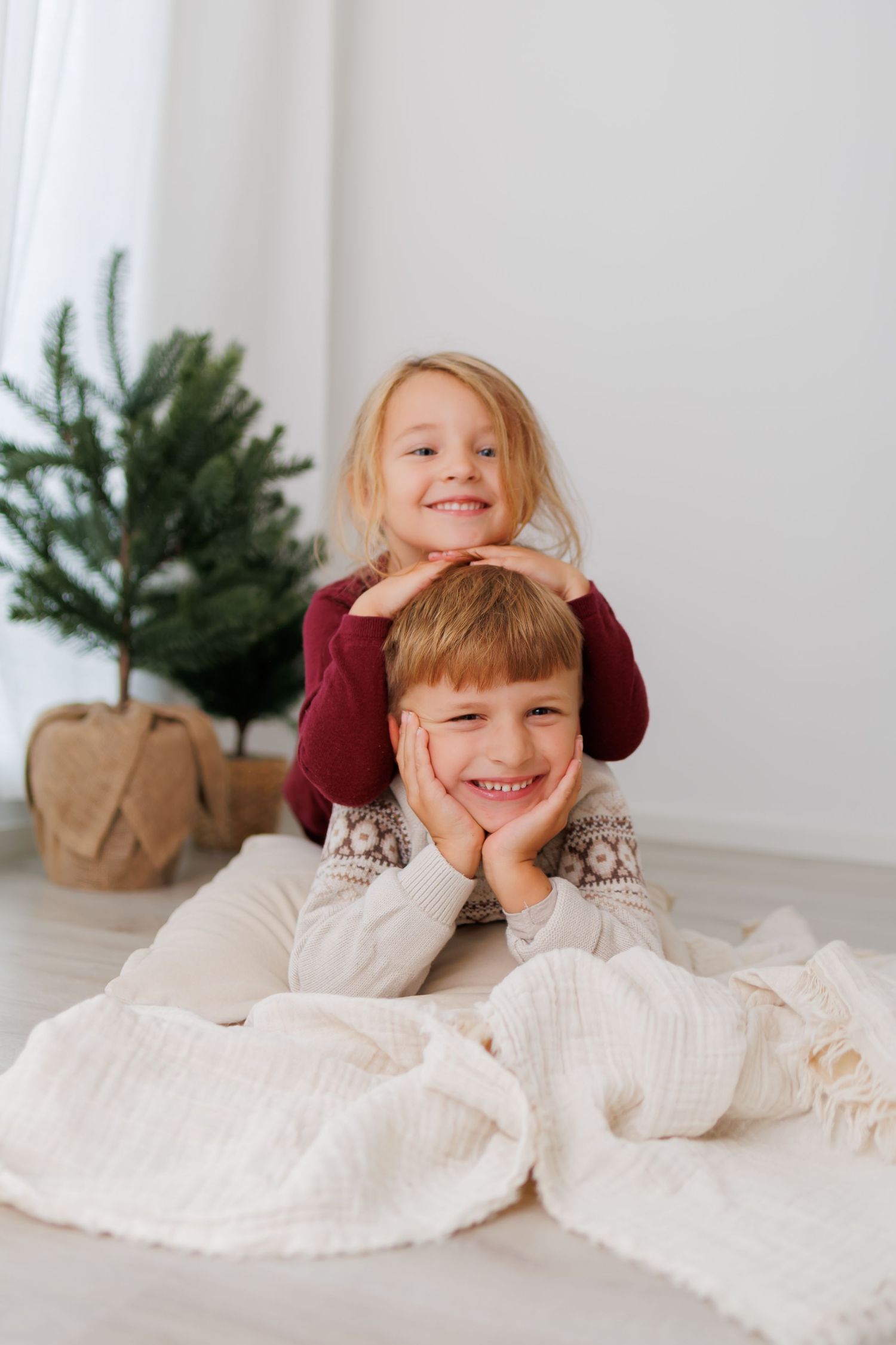 Two happy children pose together on the floor near small Christmas trees in a bright, cozy holiday setting.