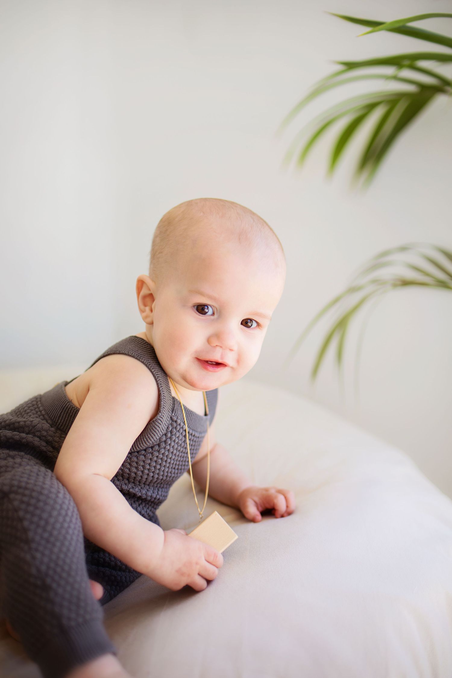 Cute baby in gray romper sitting on white sofa, smiling at camera with green plant in background.