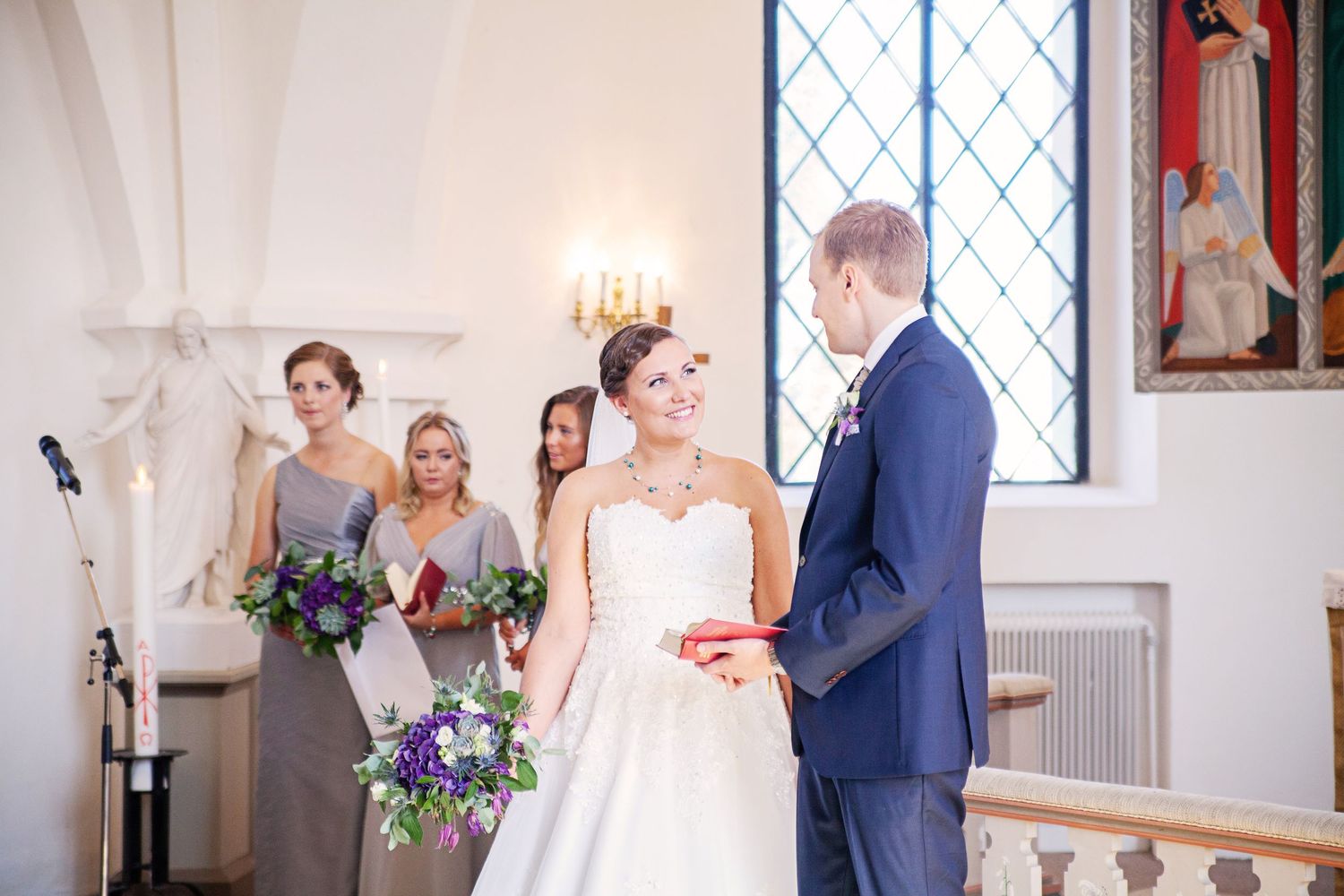 Bride and groom exchange vows at church altar with bridesmaids in grey dresses holding purple bouquets.