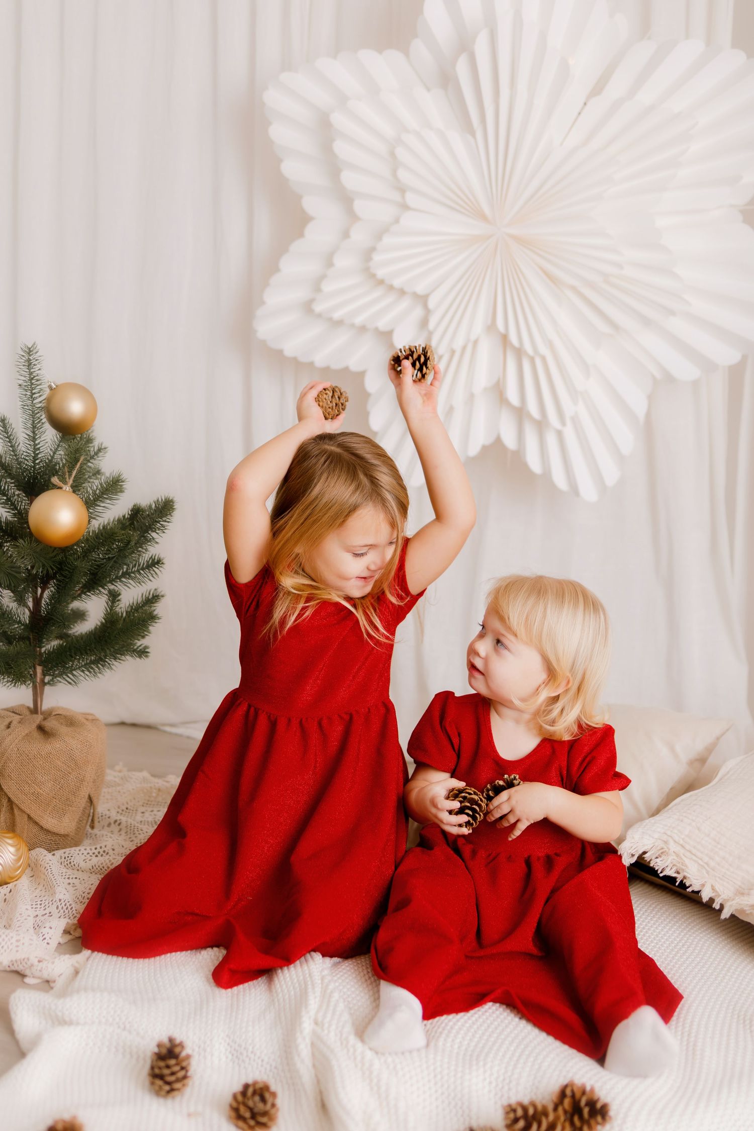 Two young girls in matching red dresses play with pine cones near a decorated Christmas tree and white paper snowflake backdrop.