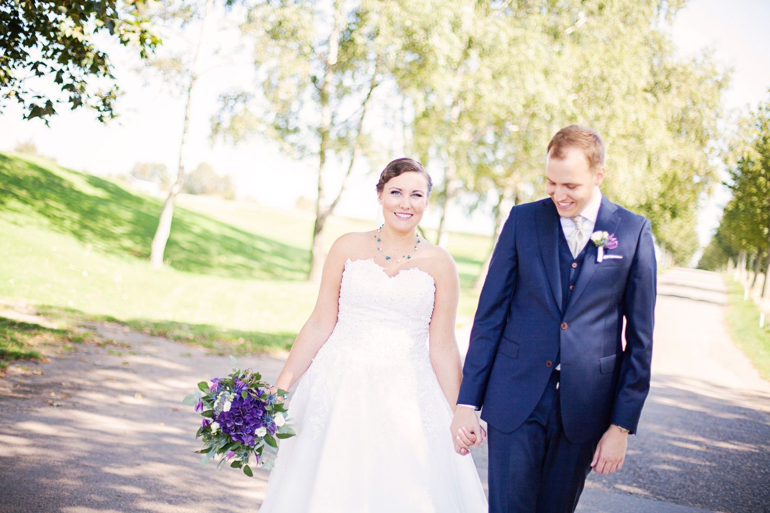 Bride and groom walking hand in hand outdoors, bride holding purple bouquet, groom in navy suit on sunny wedding day.