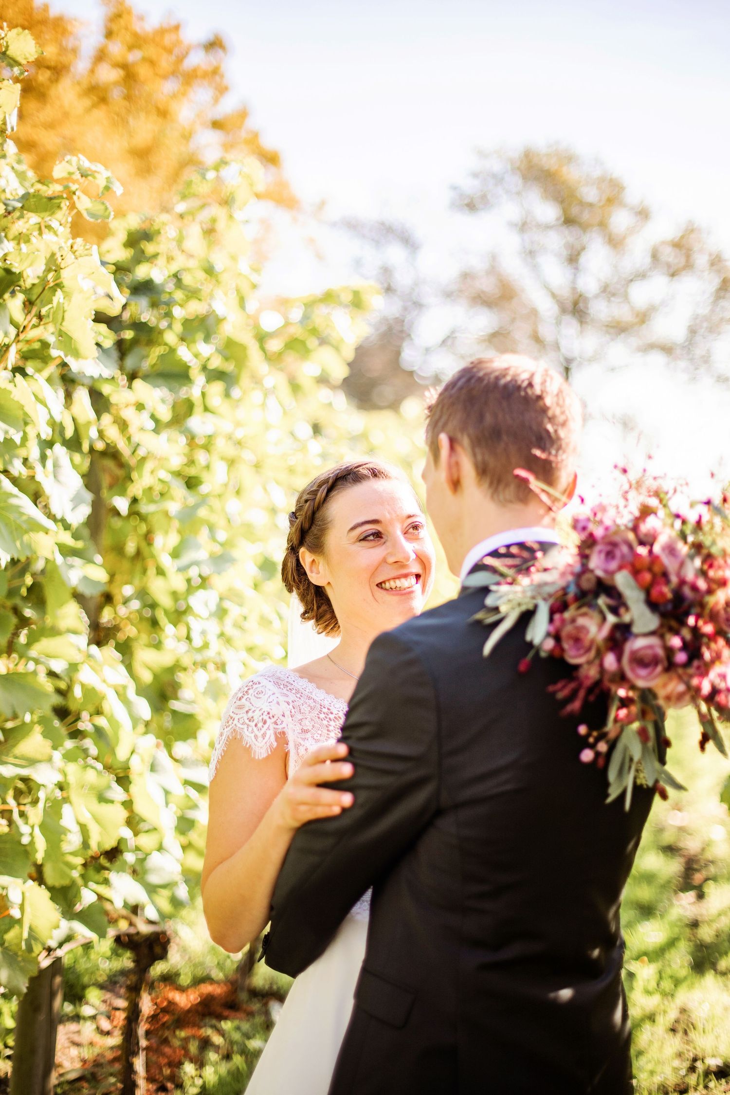 Smiling bride in white lace dress embraces groom in black suit outdoors, holding vibrant pink floral bouquet near vineyard.
