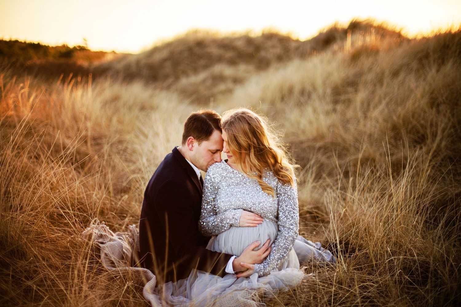 A couple sits together in golden tall grass during sunset while sharing an intimate moment.