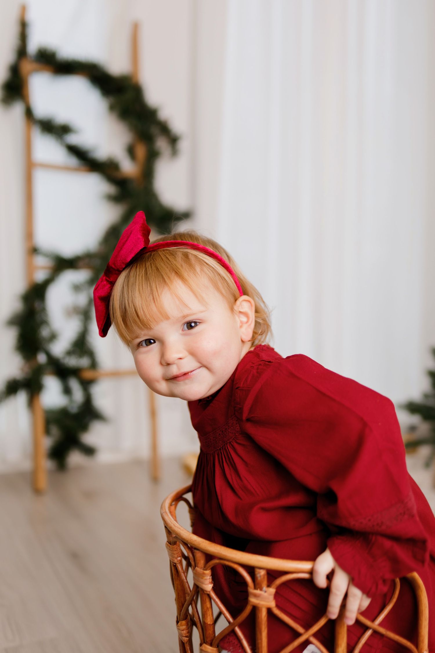 Smiling toddler girl in red dress and bow headband sitting in wicker basket with Christmas greenery decor.