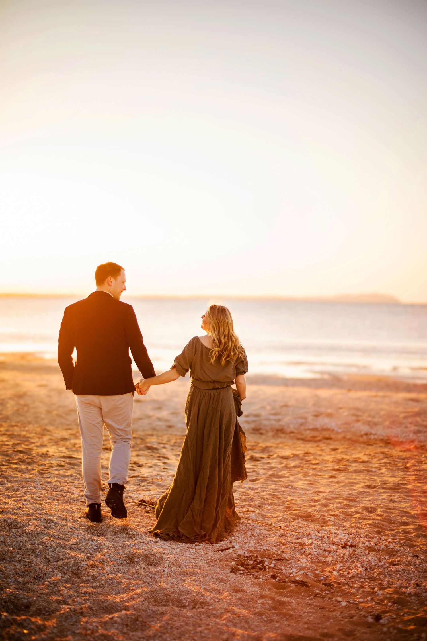A couple walks together on a sandy beach at sunset creating silhouettes against the warm golden sky.