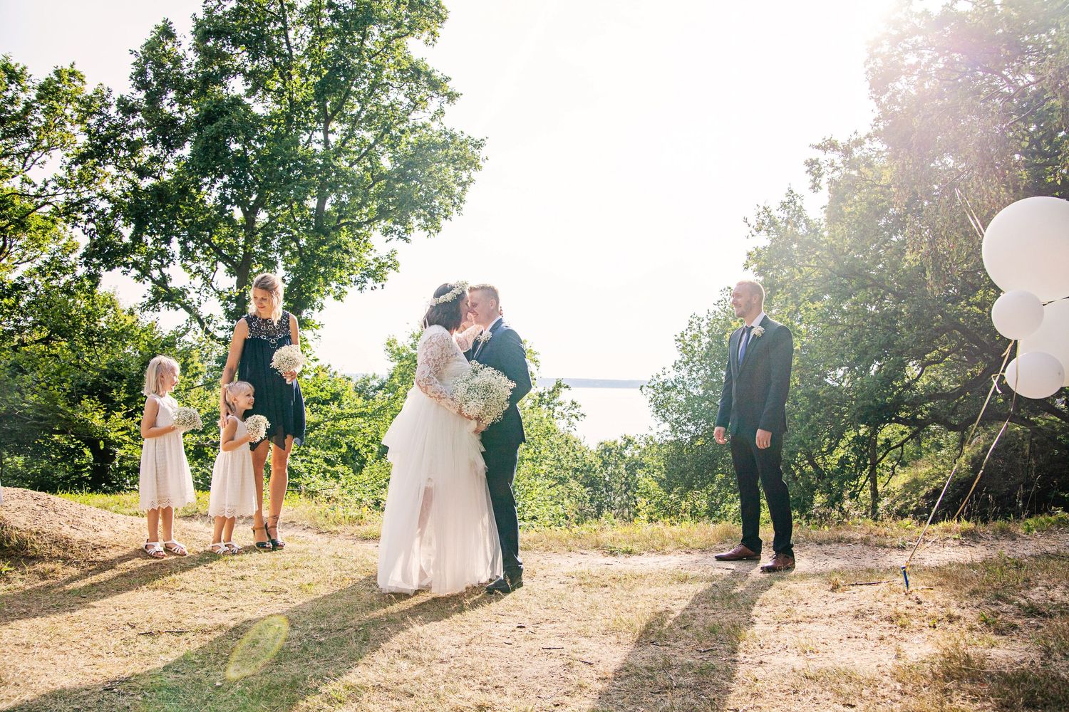 Bride and groom share a kiss outdoors at a scenic wedding ceremony with guests and white balloons nearby.