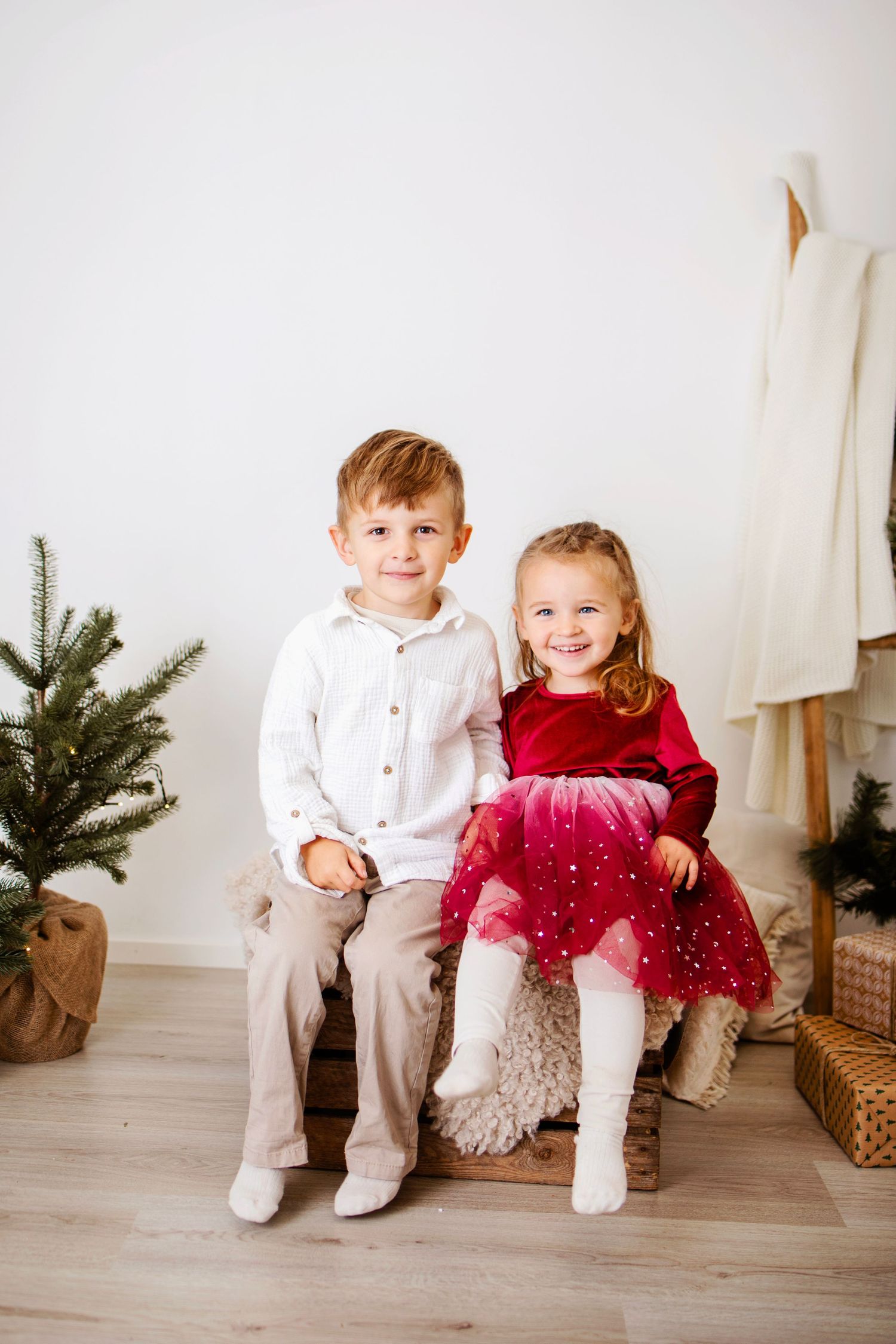 Two young children in holiday attire sitting on a bench with a small Christmas tree and wicker basket nearby.