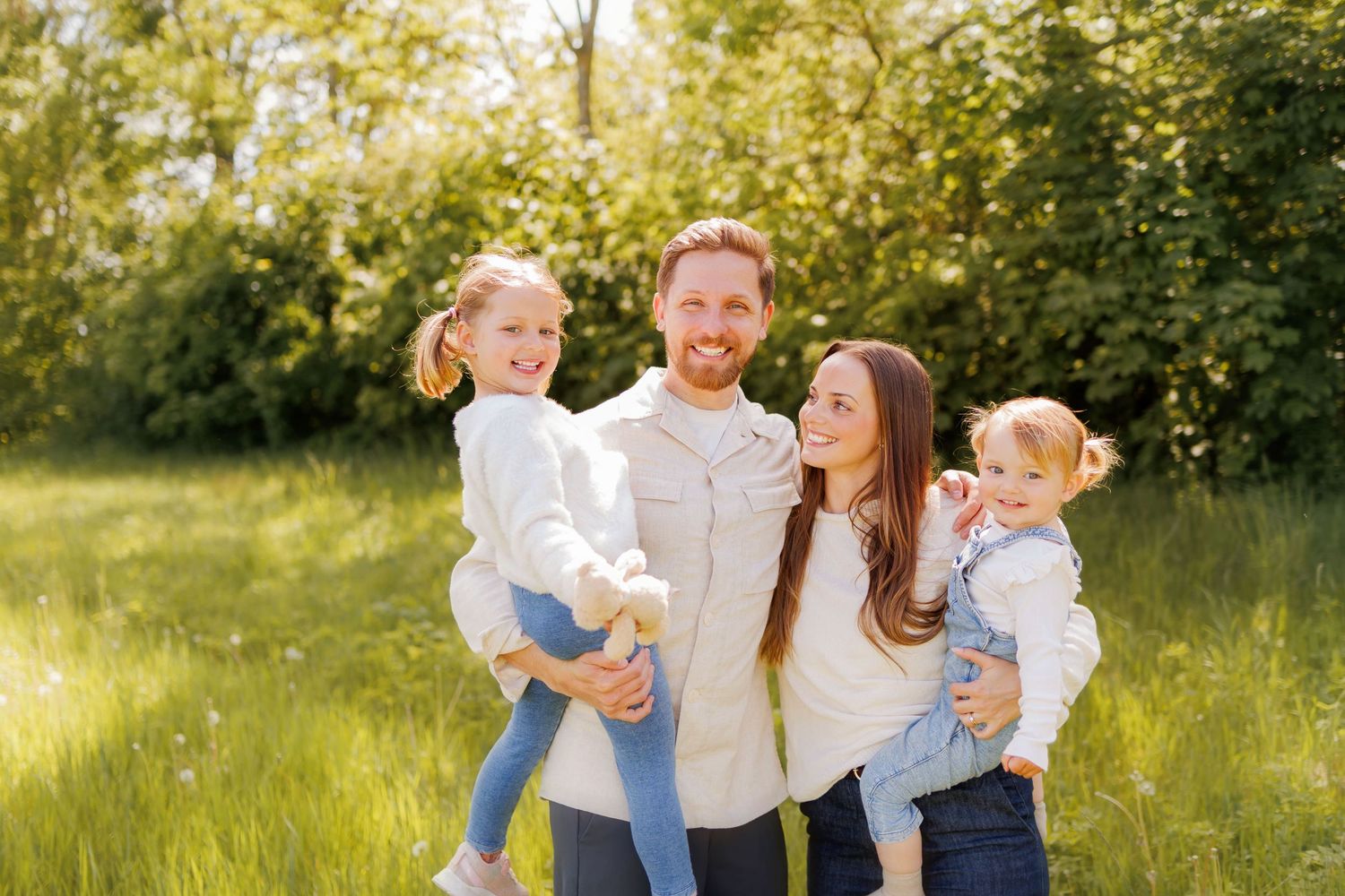 Happy family of four smiling outdoors in a sunny green meadow, parents holding two young daughters.