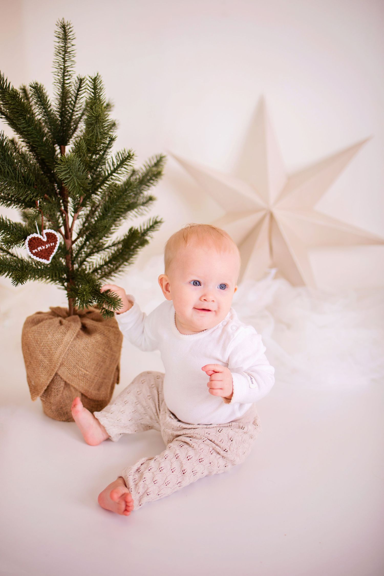 Small child in white sitting next to potted Christmas tree with heart ornament and white star decoration on wall.